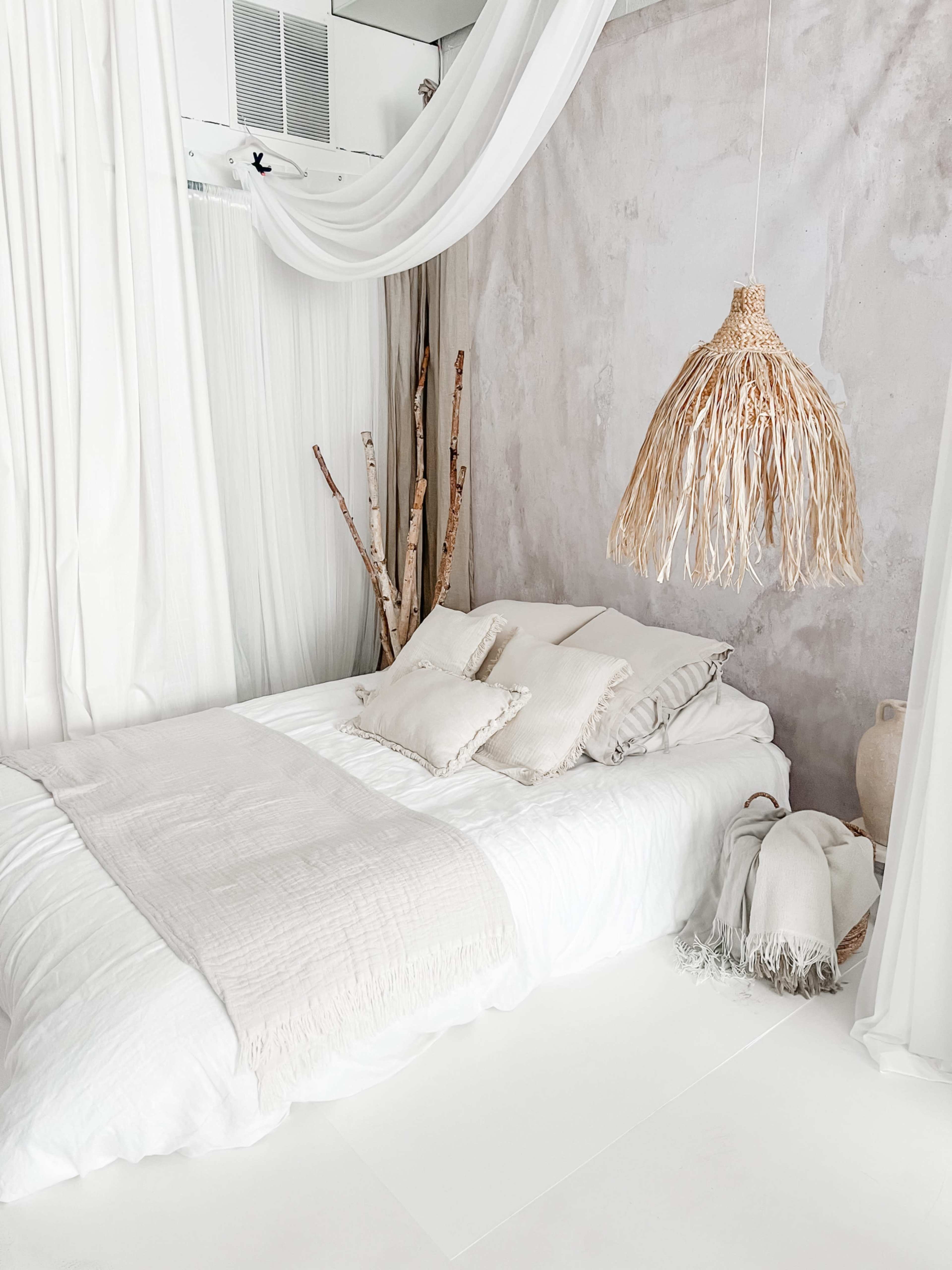 A bed with white linens is positioned against a textured light gray wall, surrounded by flowing white curtains and adorned with a woven pendant light.