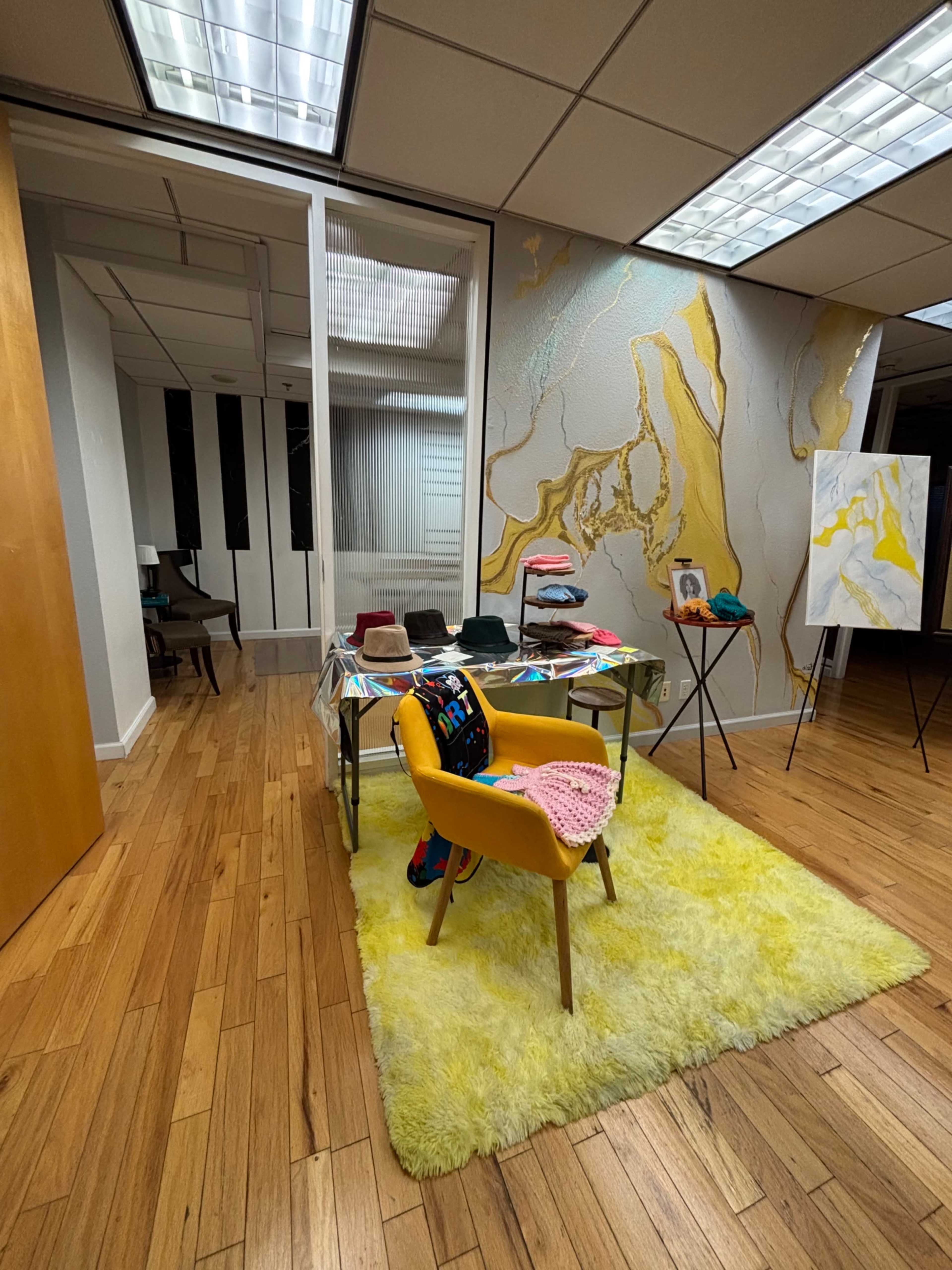 The image shows a brightly lit office space with hardwood flooring, featuring a yellow rug, a small table with clothing items, and a wall adorned with a textured gold and white design.