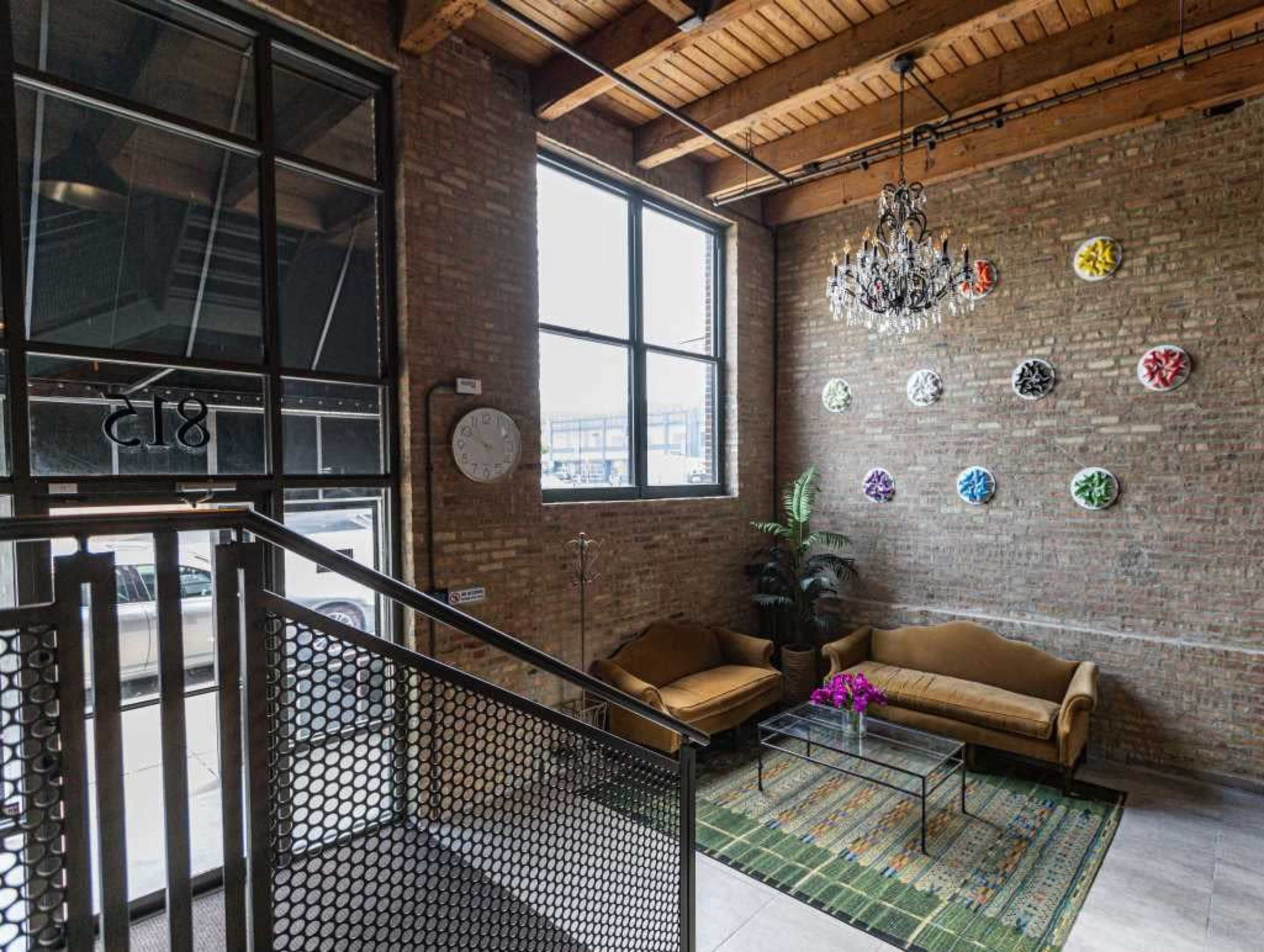The image shows a modern lobby with exposed brick walls, a chandelier, a green area rug, and two yellow sofas arranged beside a wall adorned with colorful circular artworks.