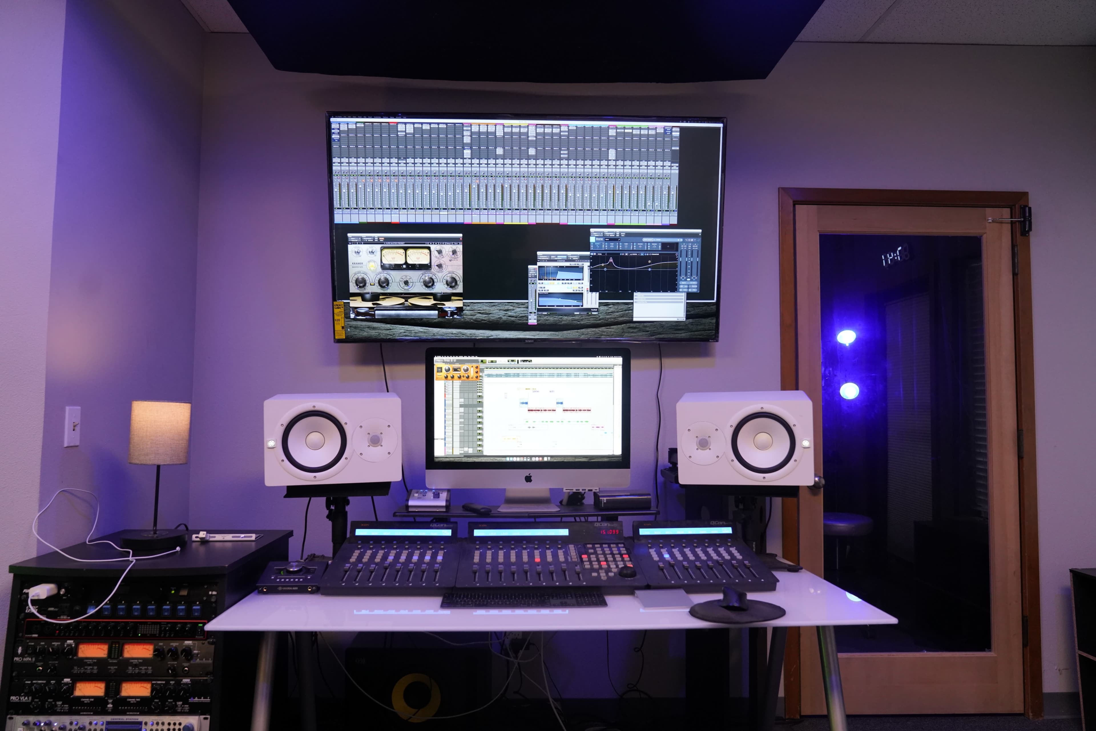 A music production studio features a large monitor displaying audio software, an iMac with mixing controls, and dual studio speakers on a desk.