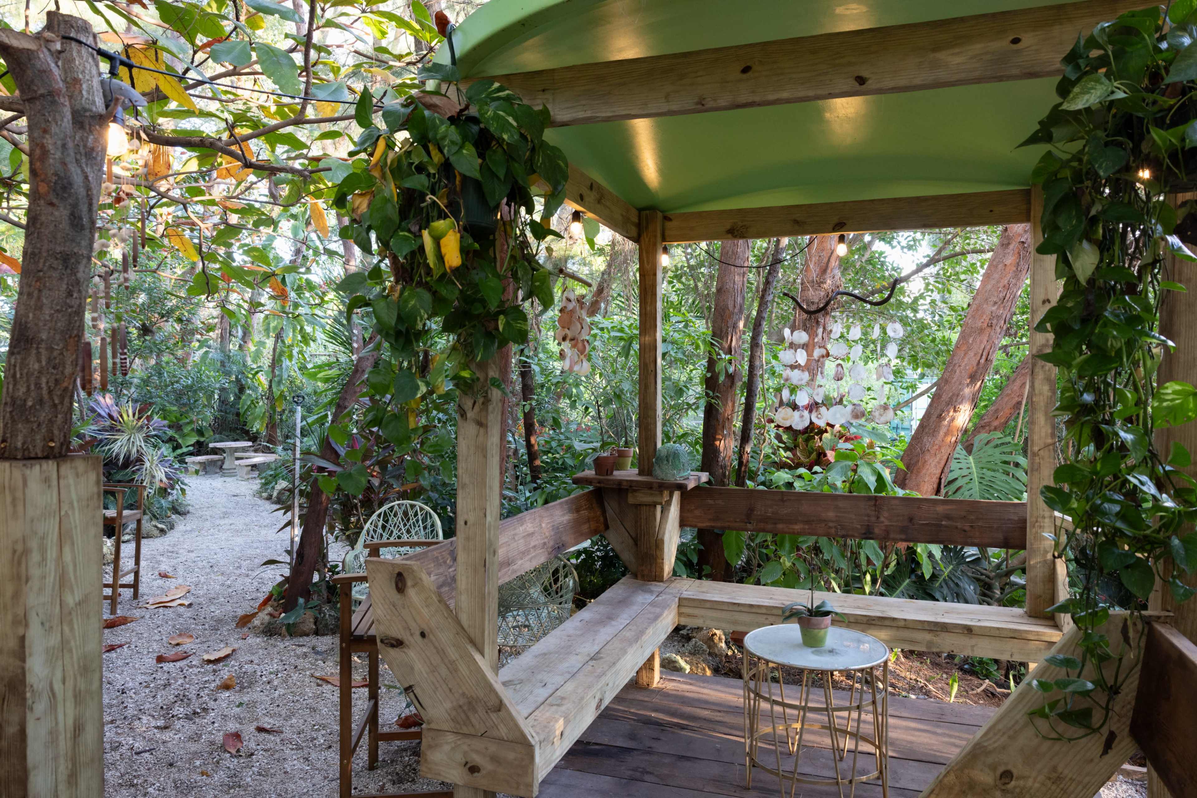 The image shows a wooden gazebo surrounded by lush greenery, with a small round table and a bench seating area.
