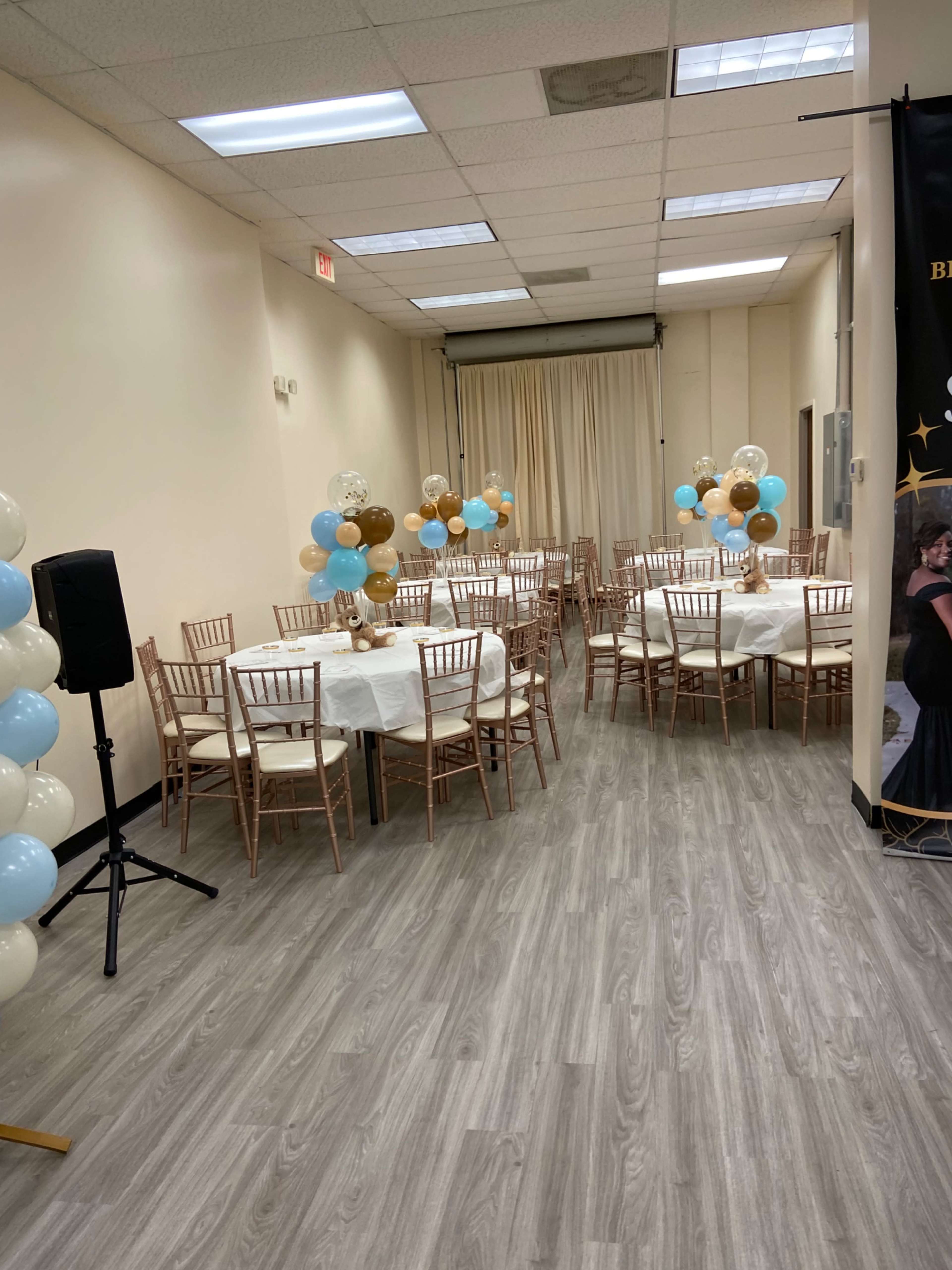 The image shows a decorated event space with tables set for a gathering, featuring balloons in blue and gold accents.