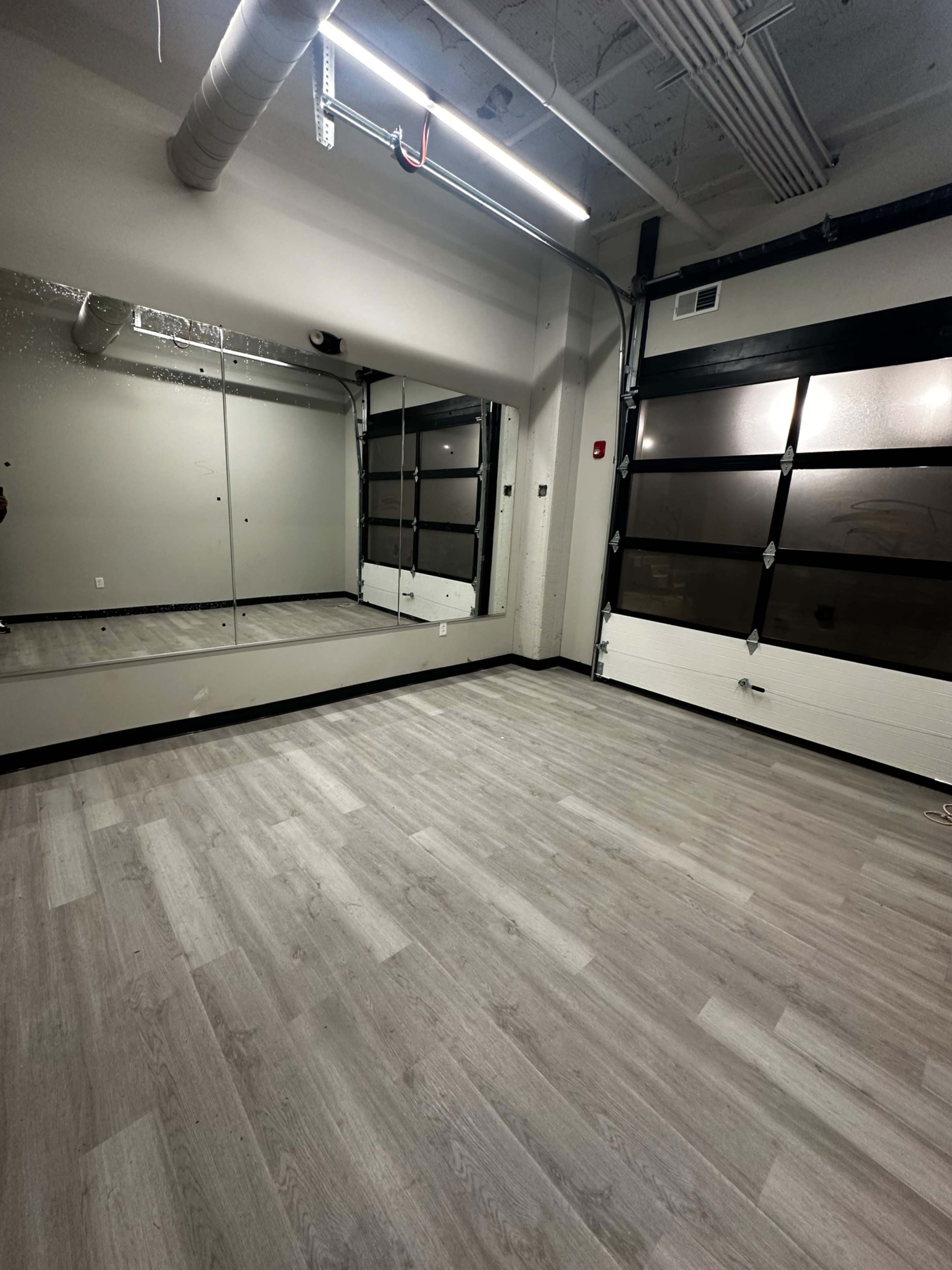 The image shows an empty room with gray wooden floors, large mirrors on one wall, and a garage-style door partially visible on the opposite wall.