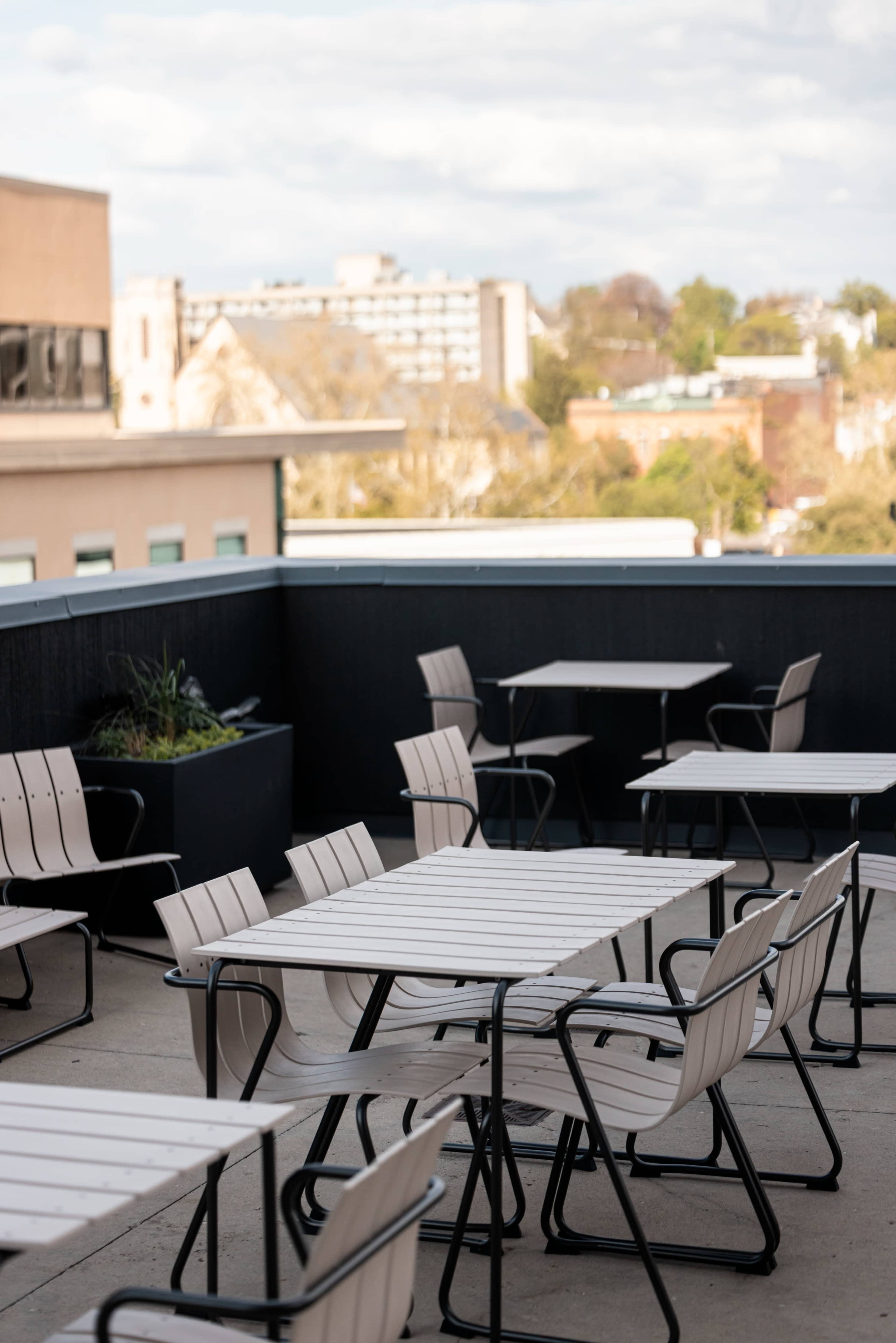 The image shows a rooftop terrace with empty wooden tables and chairs, overlooking a cityscape in the background.