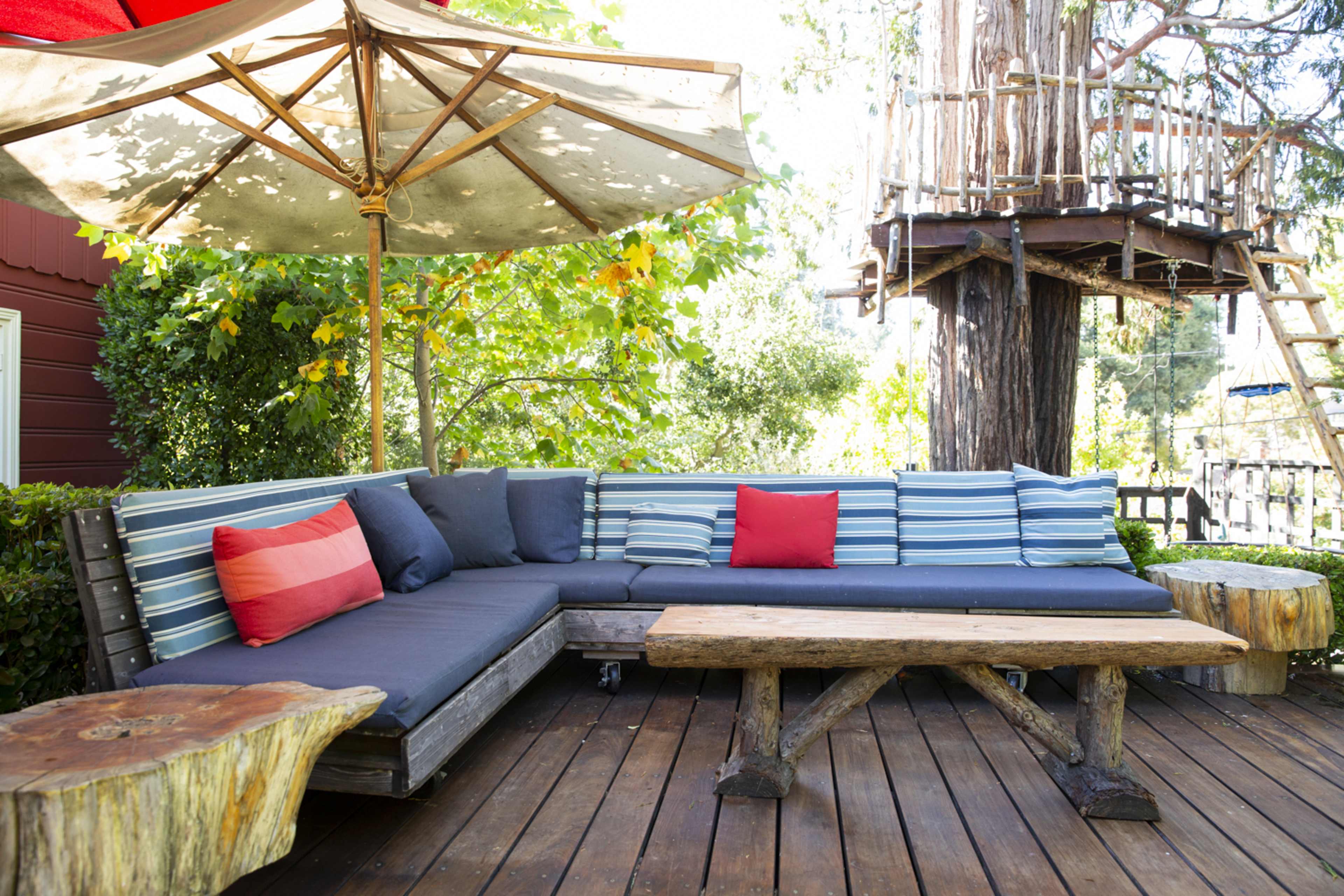 A wooden deck features a sectional sofa with striped cushions, a wooden table, and a large umbrella shaded by trees.