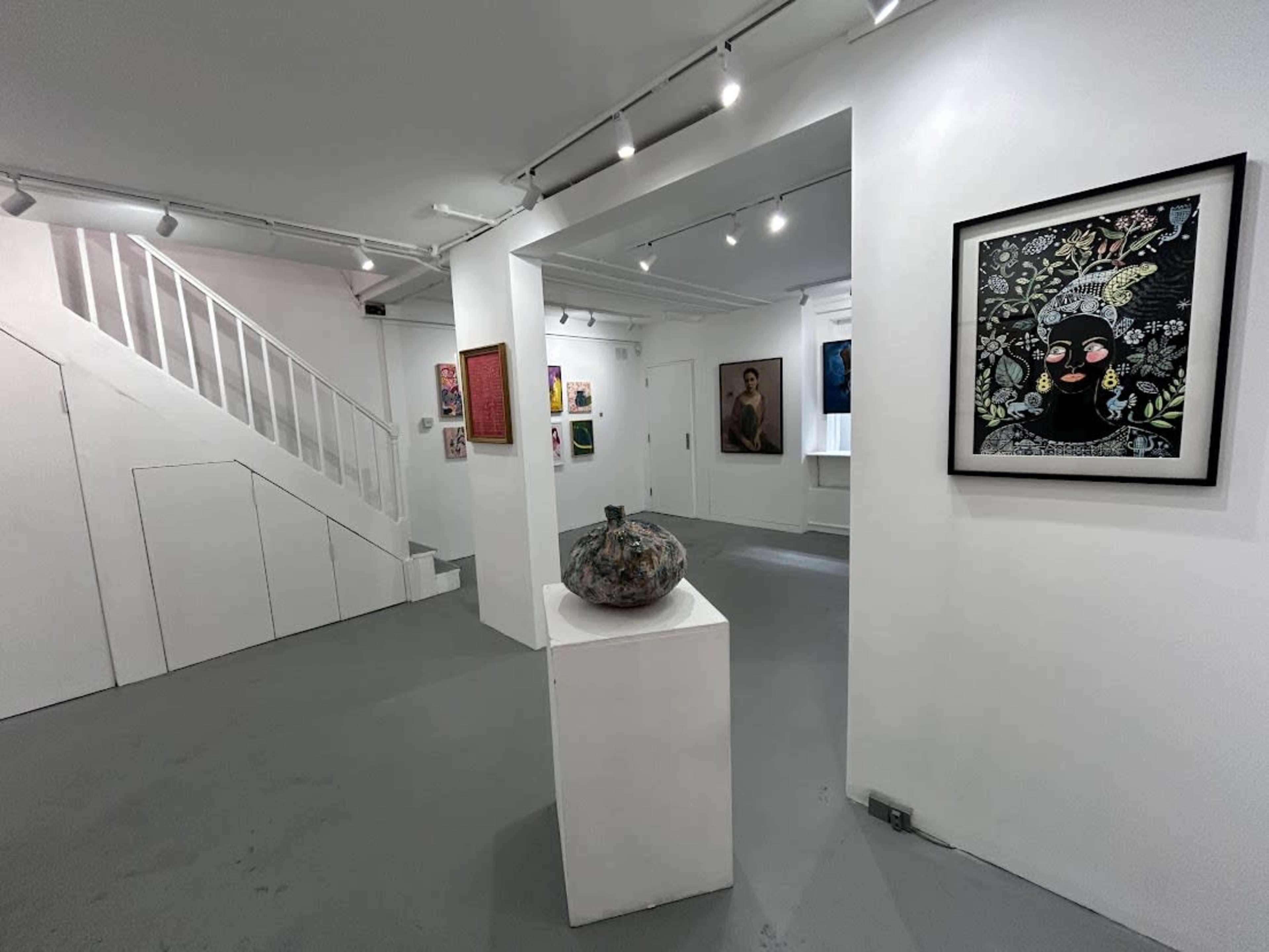 An art gallery with a white interior featuring various artworks on the walls, a sculpture on a pedestal, and a staircase in the background.