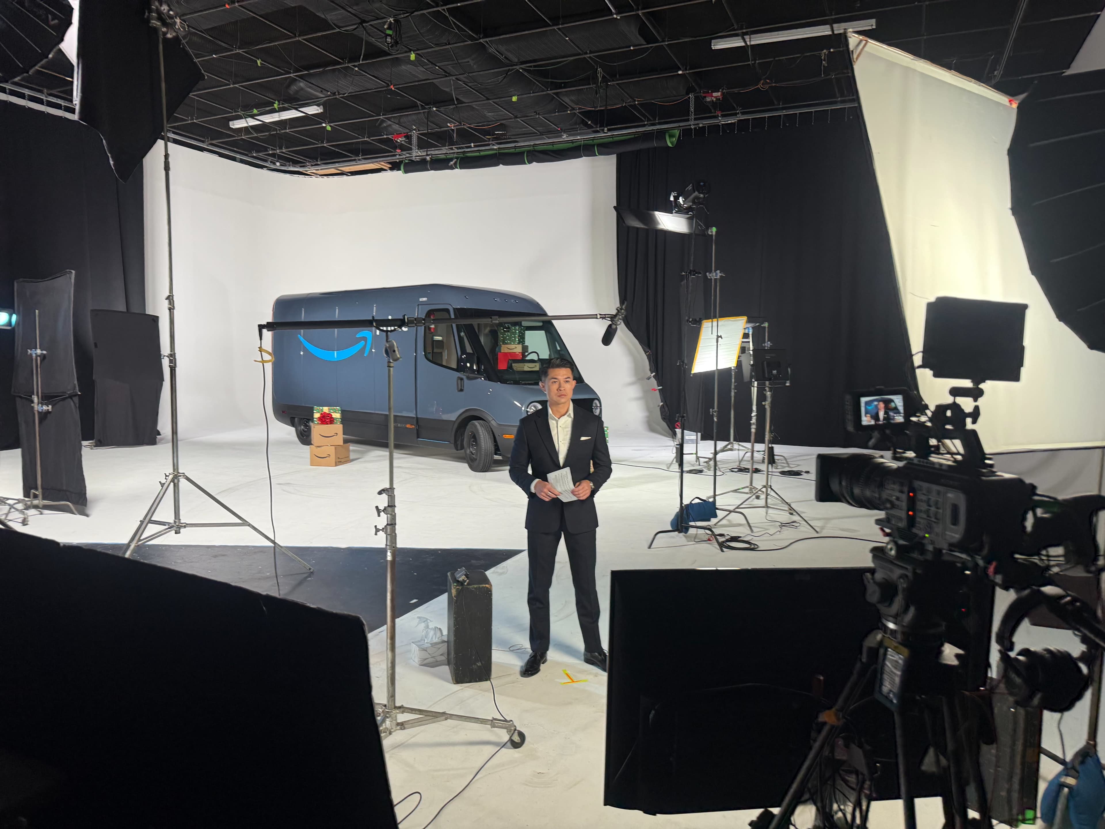 A man in a suit stands in a studio in front of an Amazon delivery van, with cameras and lighting equipment surrounding him.