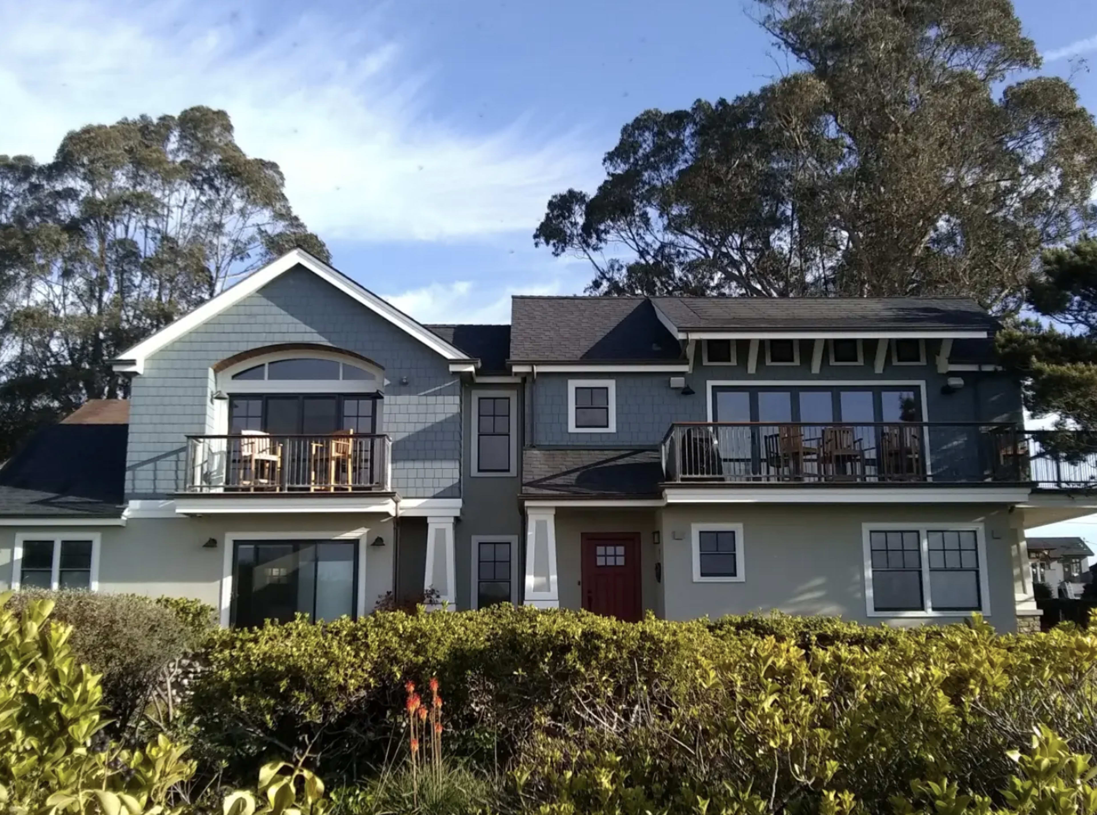 The image shows a two-story blue house with balconies and a red front door, surrounded by green shrubs and trees.