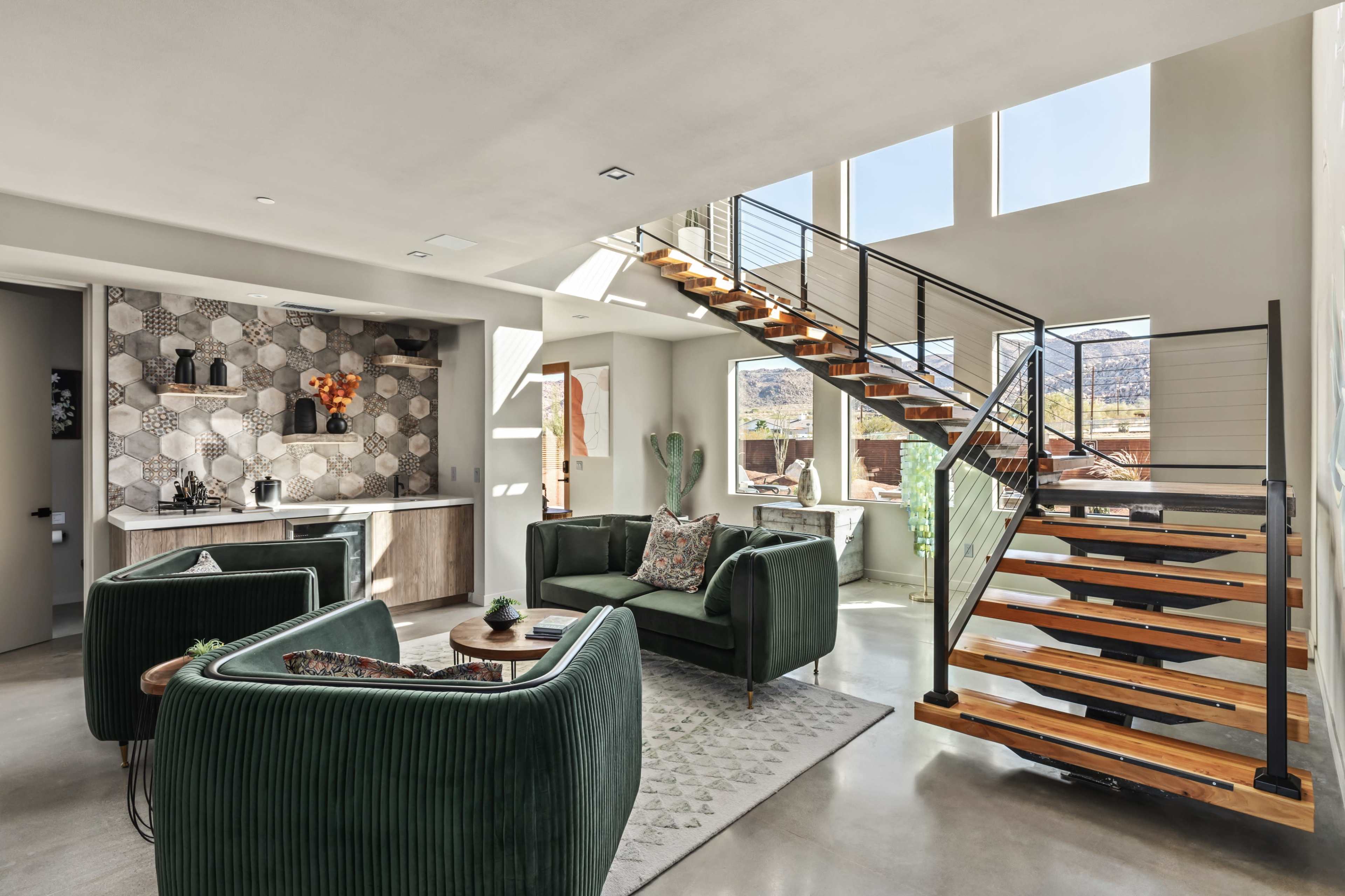 The image shows a modern living space with green velvet armchairs, a staircase with wooden steps, and large windows that allow natural light to fill the room.