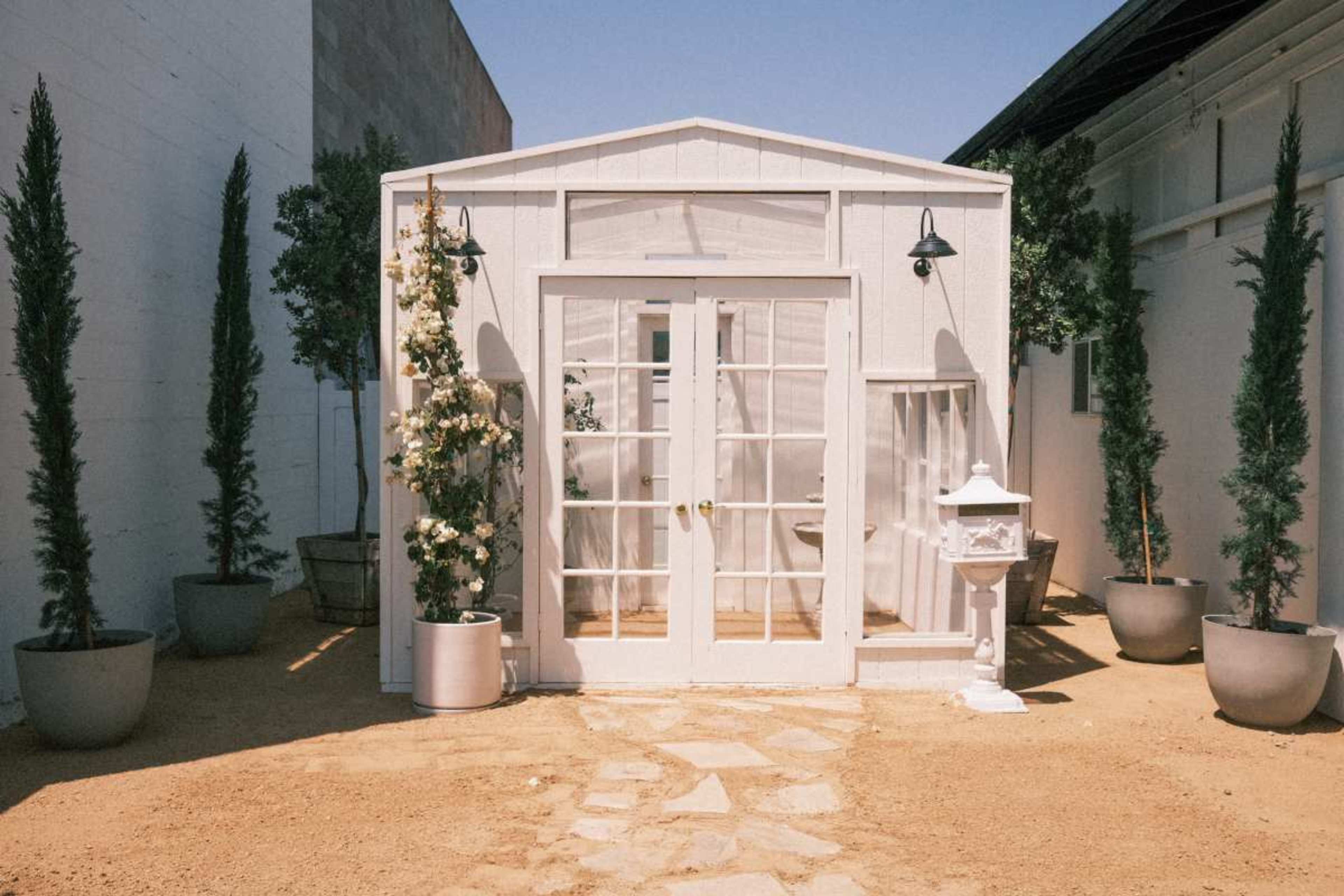 A white garden shed with clear glass doors is surrounded by potted plants and a gravel pathway.