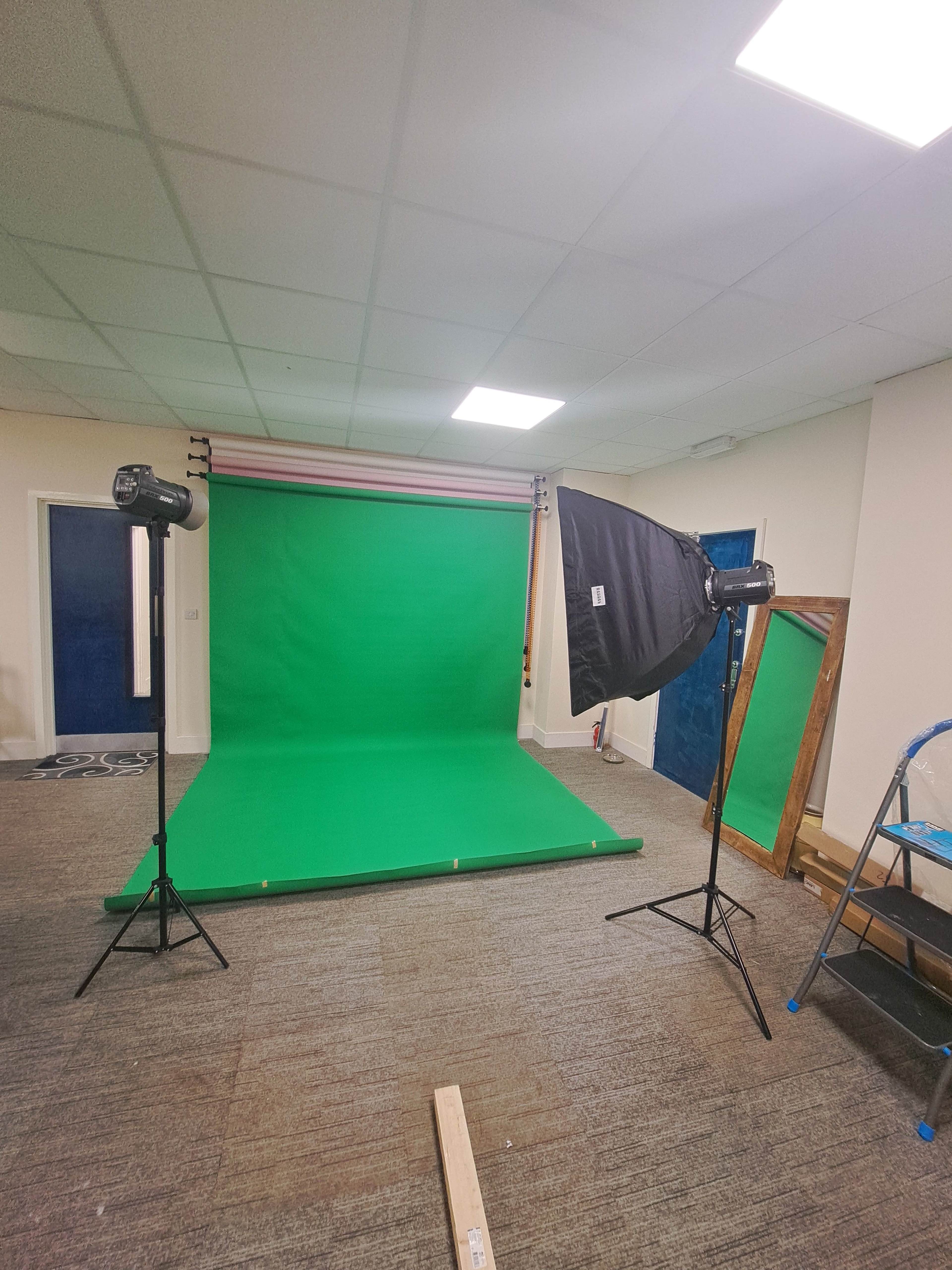 A photography setup featuring a green backdrop, two lighting softboxes on tripods, and a wooden mirror against the wall in a well-lit room.