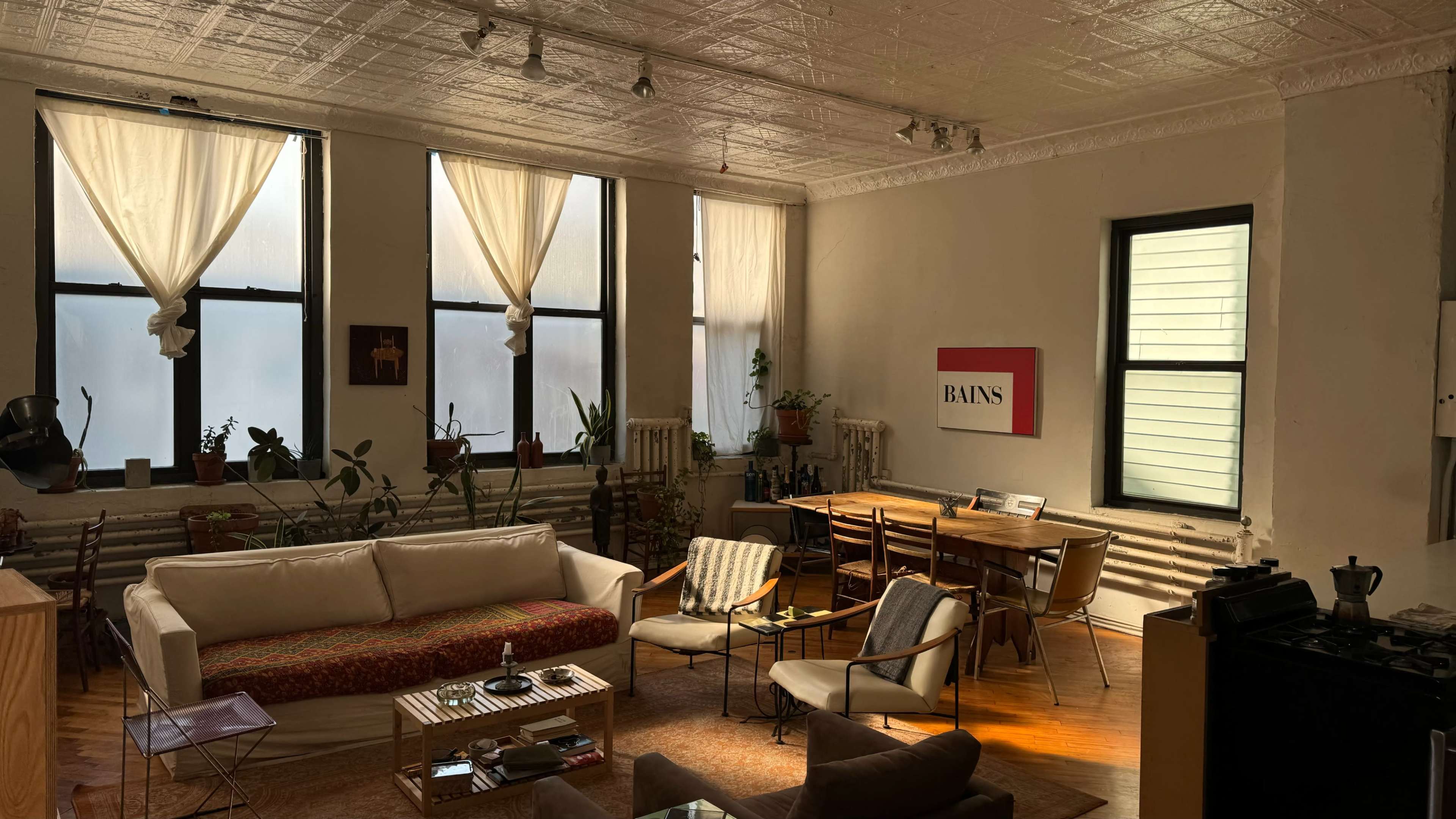A bright living room features large windows with sheer curtains, a white couch, a dining area with wooden chairs, and various plants scattered throughout the space.