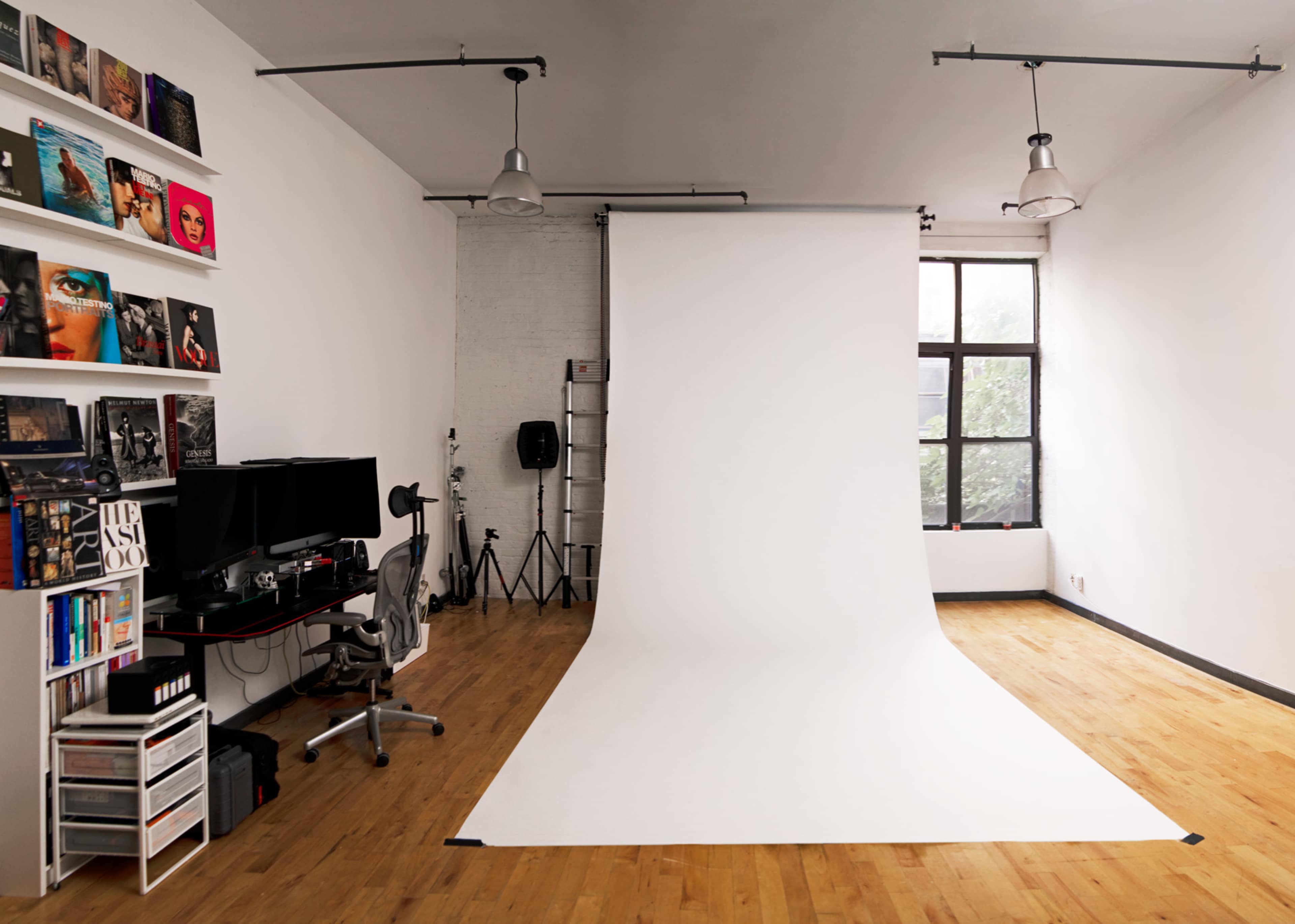 A photography studio features a white backdrop, wooden flooring, and equipment arranged near a large window.