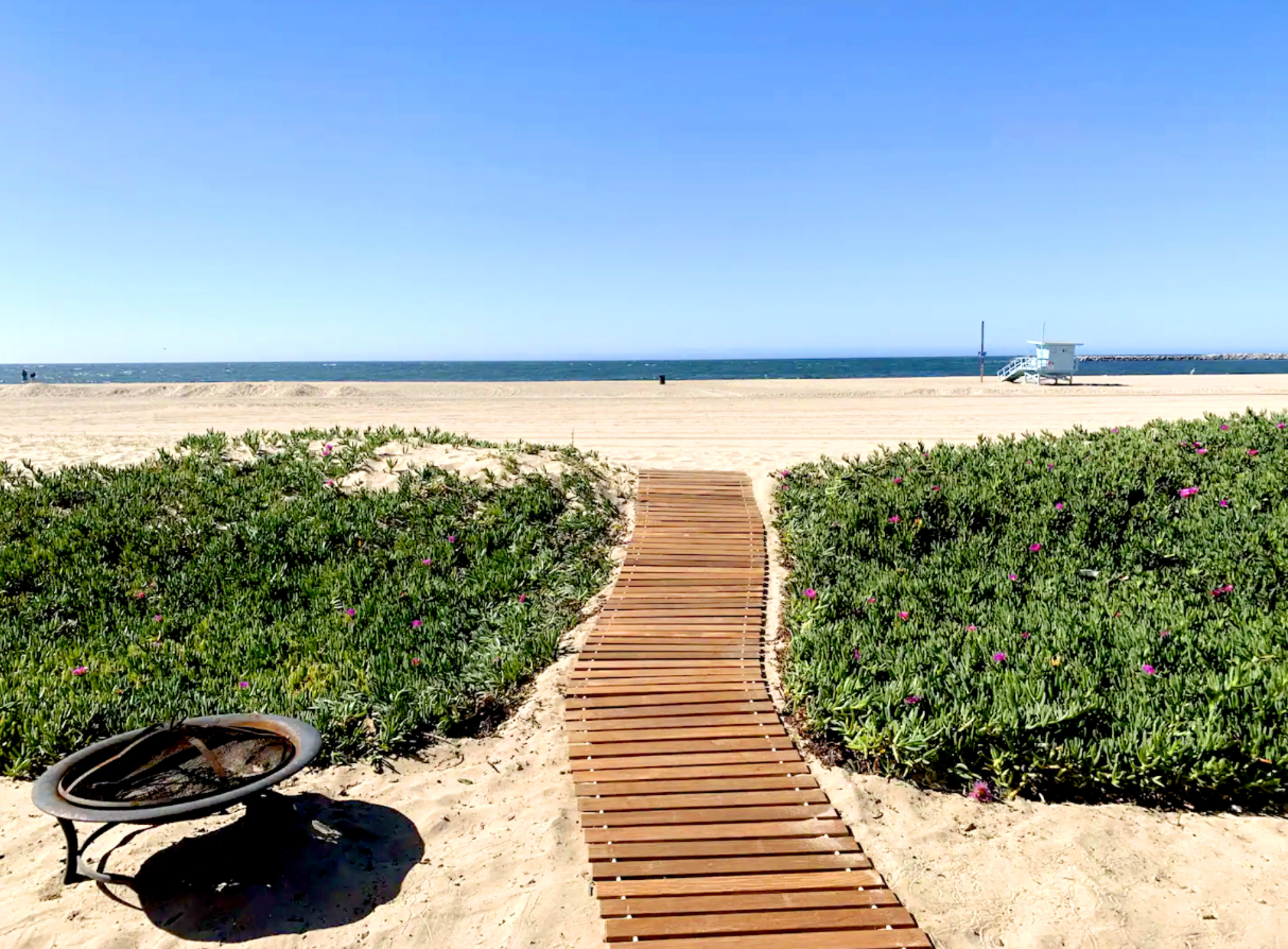 A wooden pathway leads through sand and vegetation to the ocean's edge, with a lifeguard station visible in the distance.