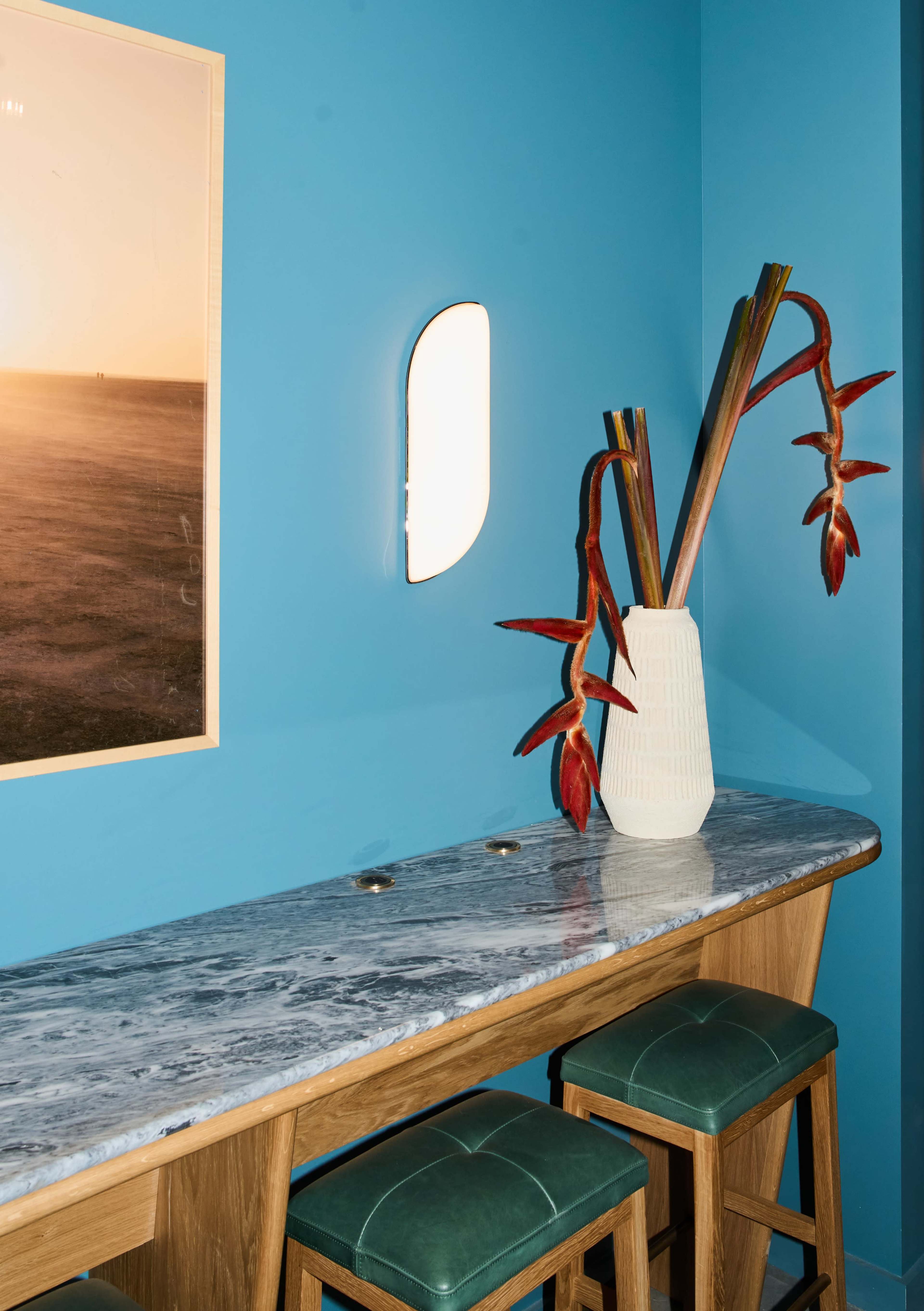 A marble-top bar with stools is positioned against a bright blue wall, accented by a large photo and a vase of decorative flowers.