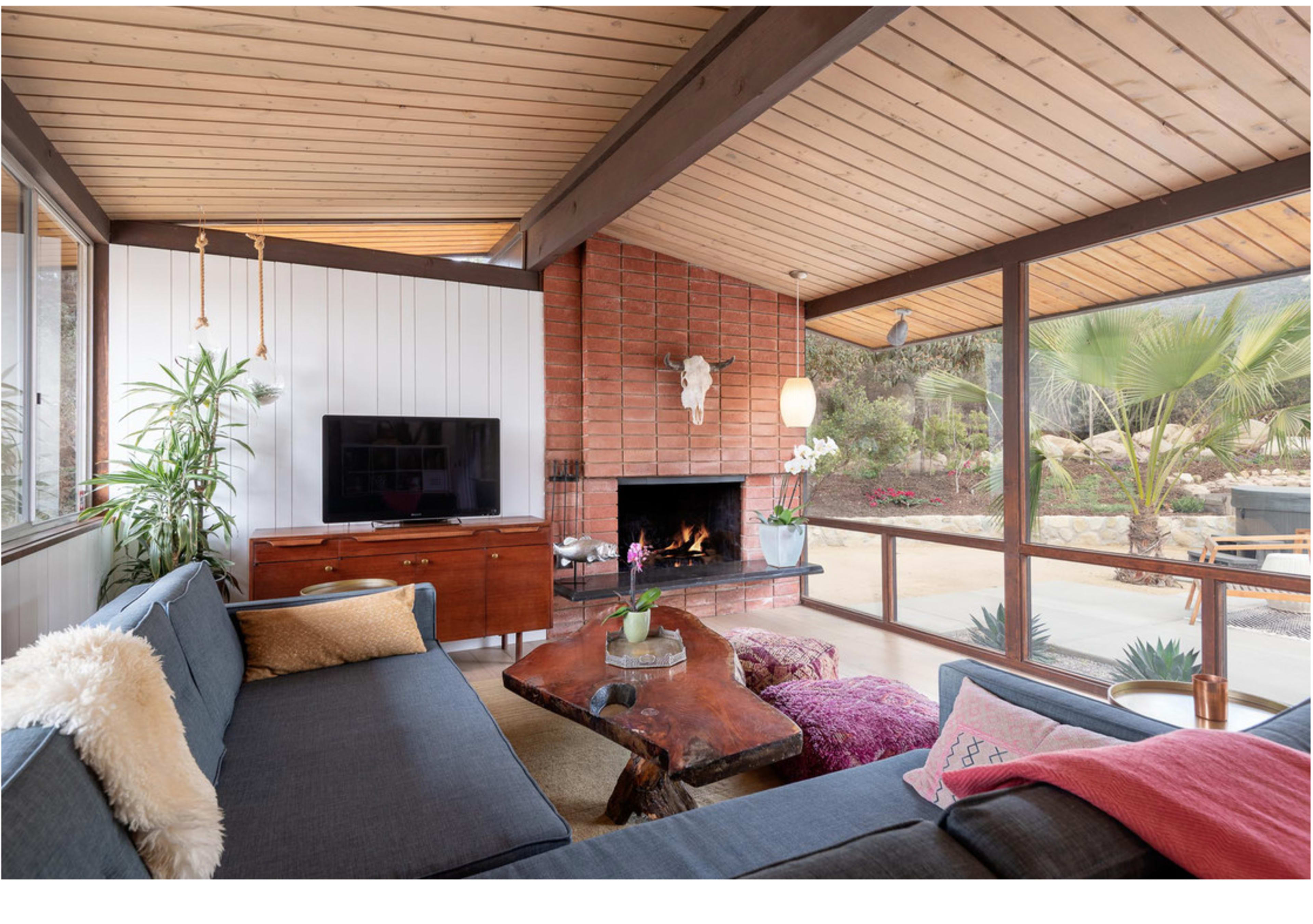 A modern living room features a wood-paneled ceiling, a large sectional sofa, a coffee table made from natural wood, and a fireplace surrounded by brick.