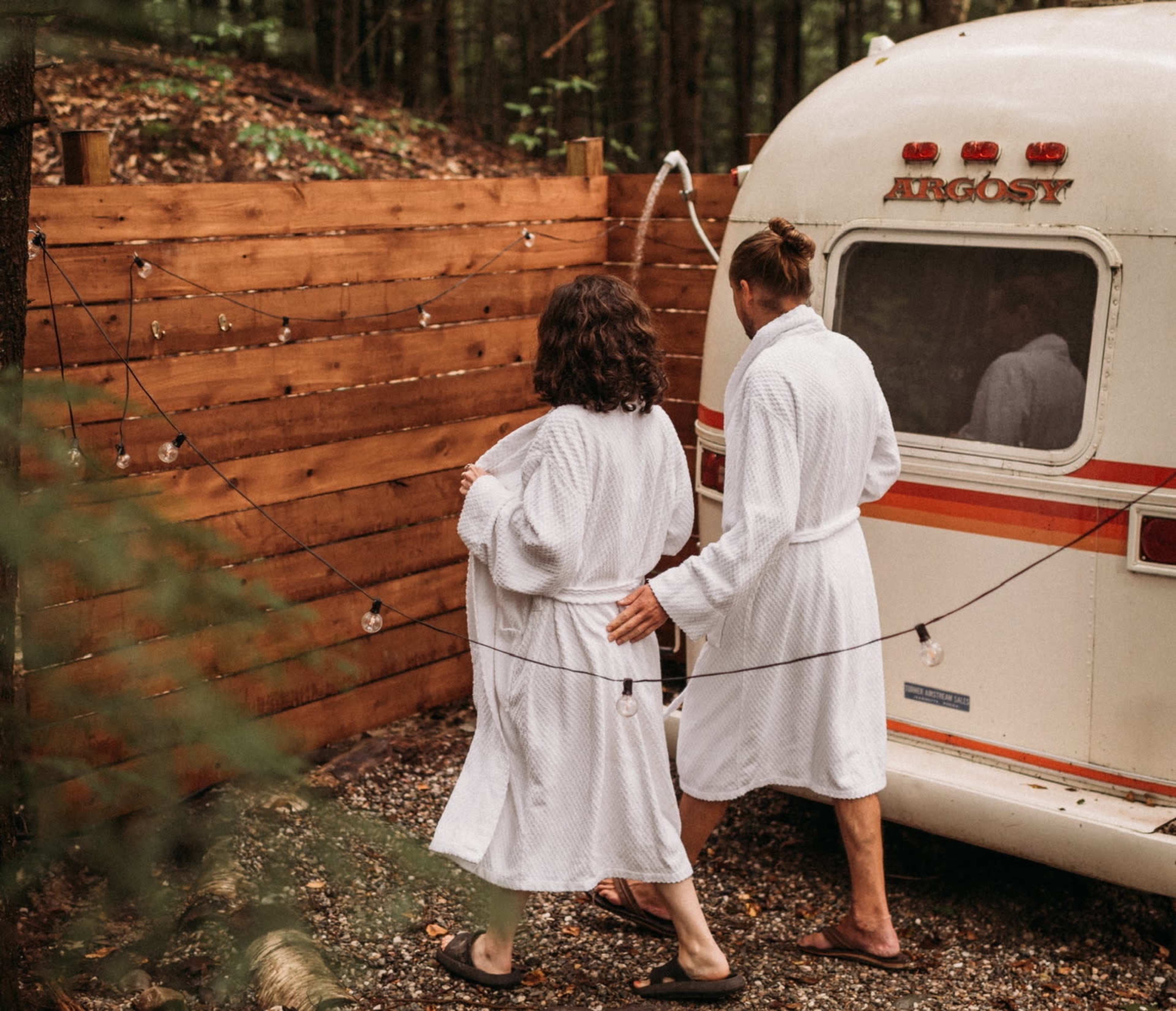 Two people in white bathrobes walk toward a vintage trailer surrounded by wooden fencing and string lights.
