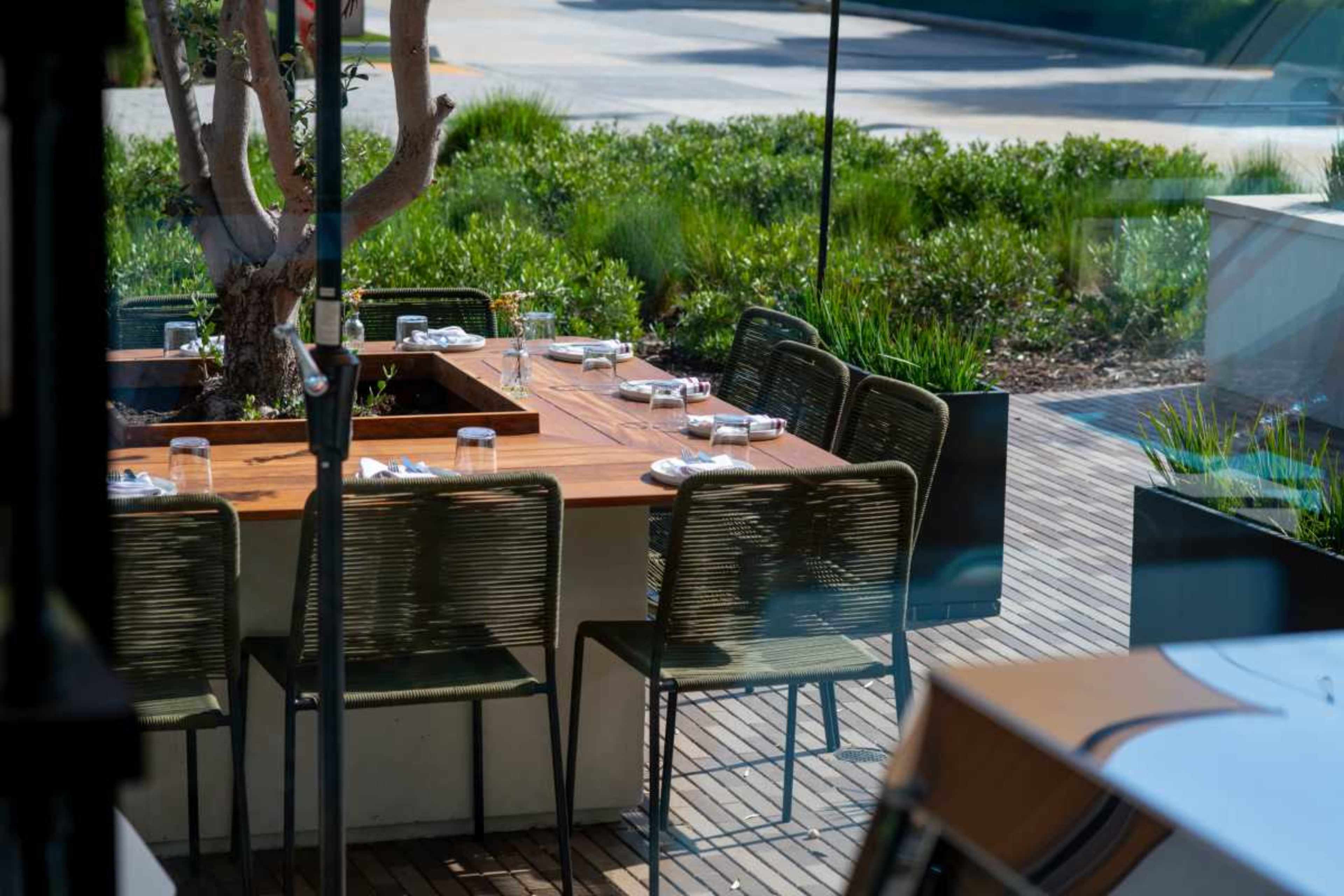 A rectangular wooden table is set with plates and glasses, surrounded by chairs on a wooden deck, with greenery in the background.
