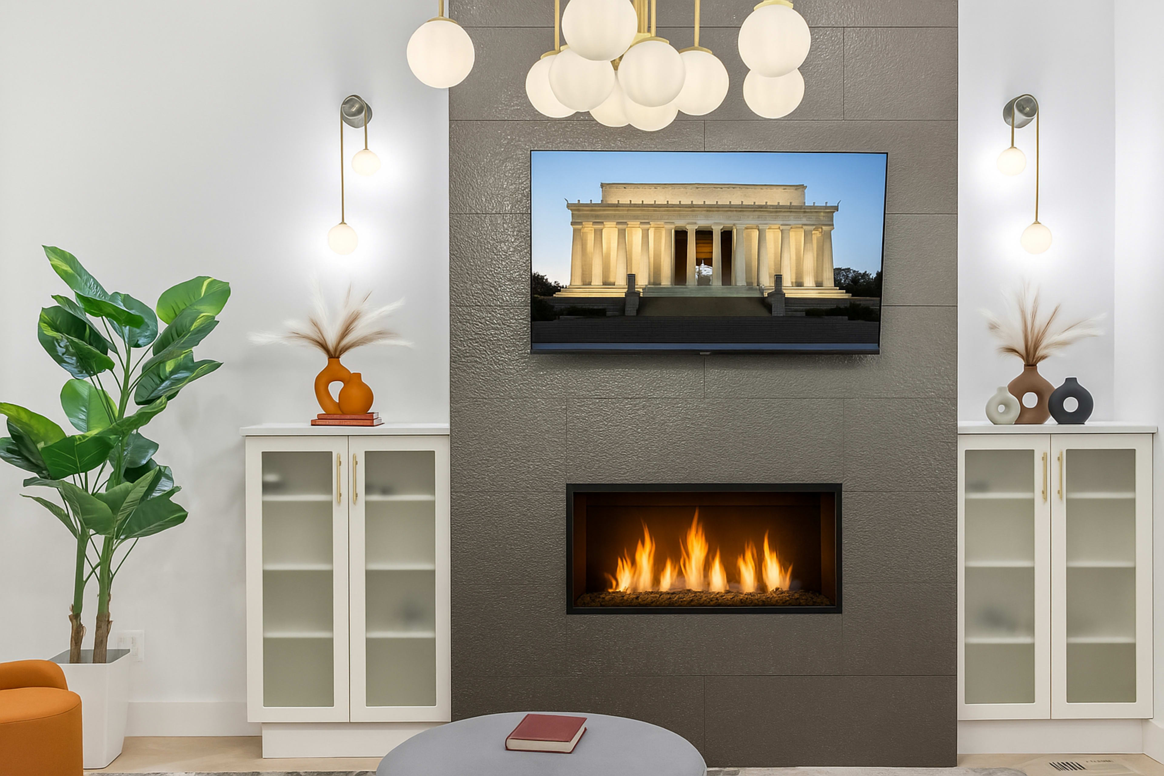 The image shows a modern living room featuring a fireplace under a mounted television displaying the Lincoln Memorial, with decorative plants and furniture arranged around the space.