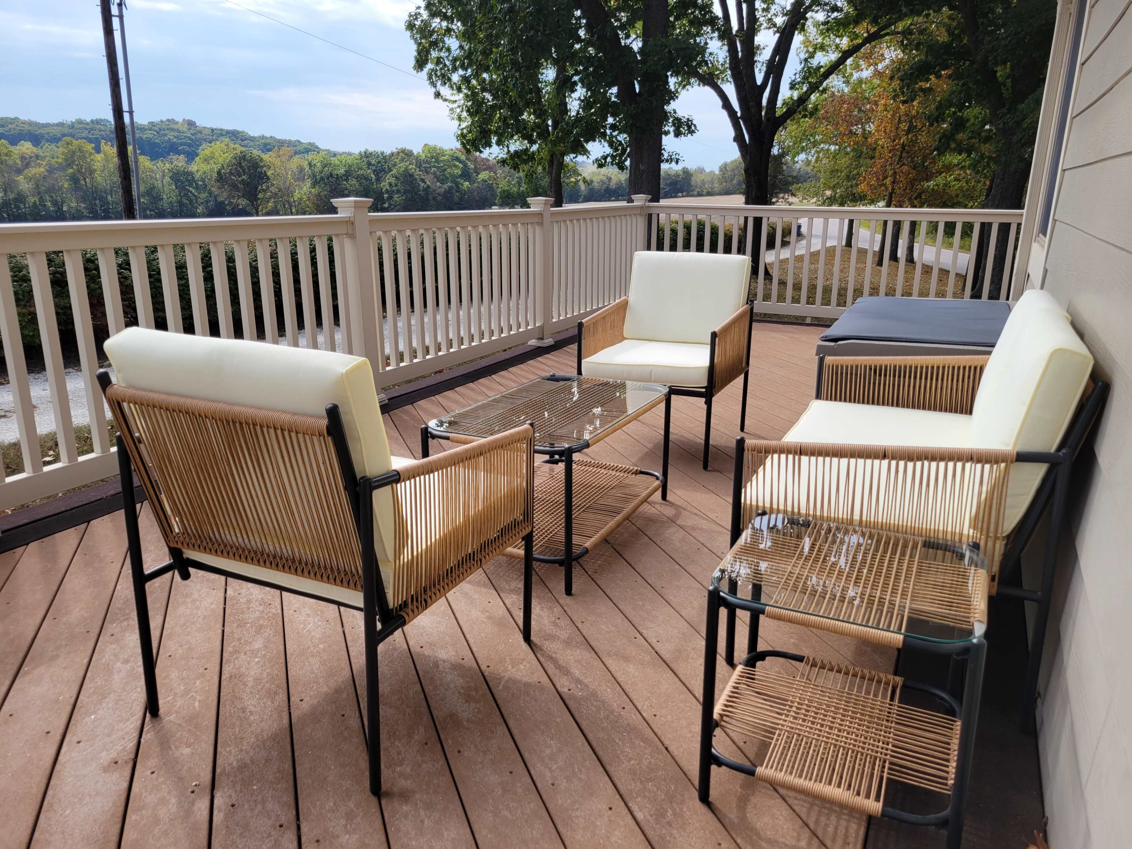 The image shows a deck with four chairs and a glass-top table arranged under a cloudy sky, surrounded by trees and a view of a distant hillside.