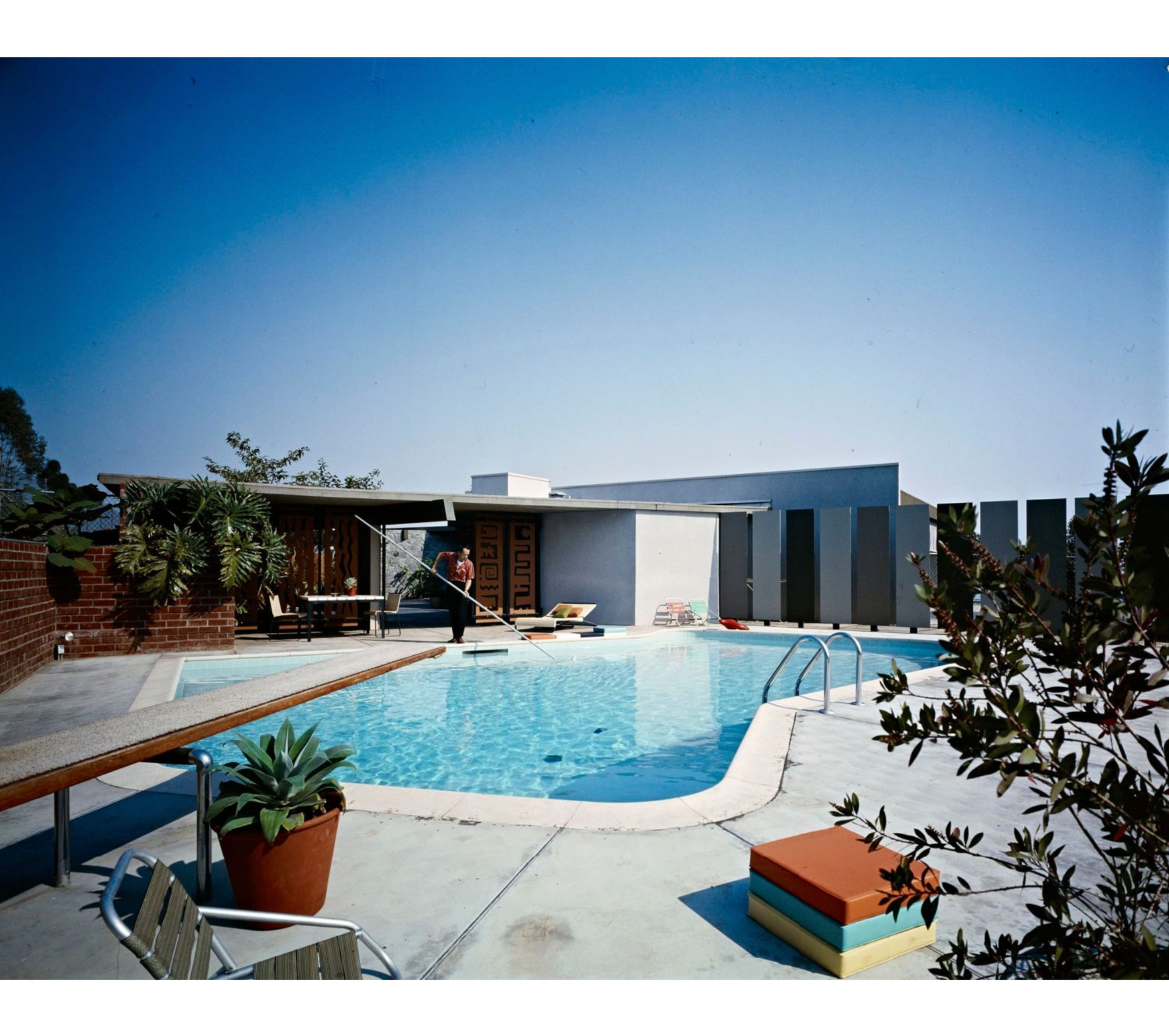 The image shows a bright, modern swimming pool area with lounge chairs, potted plants, and a backdrop of a minimalist architectural structure.