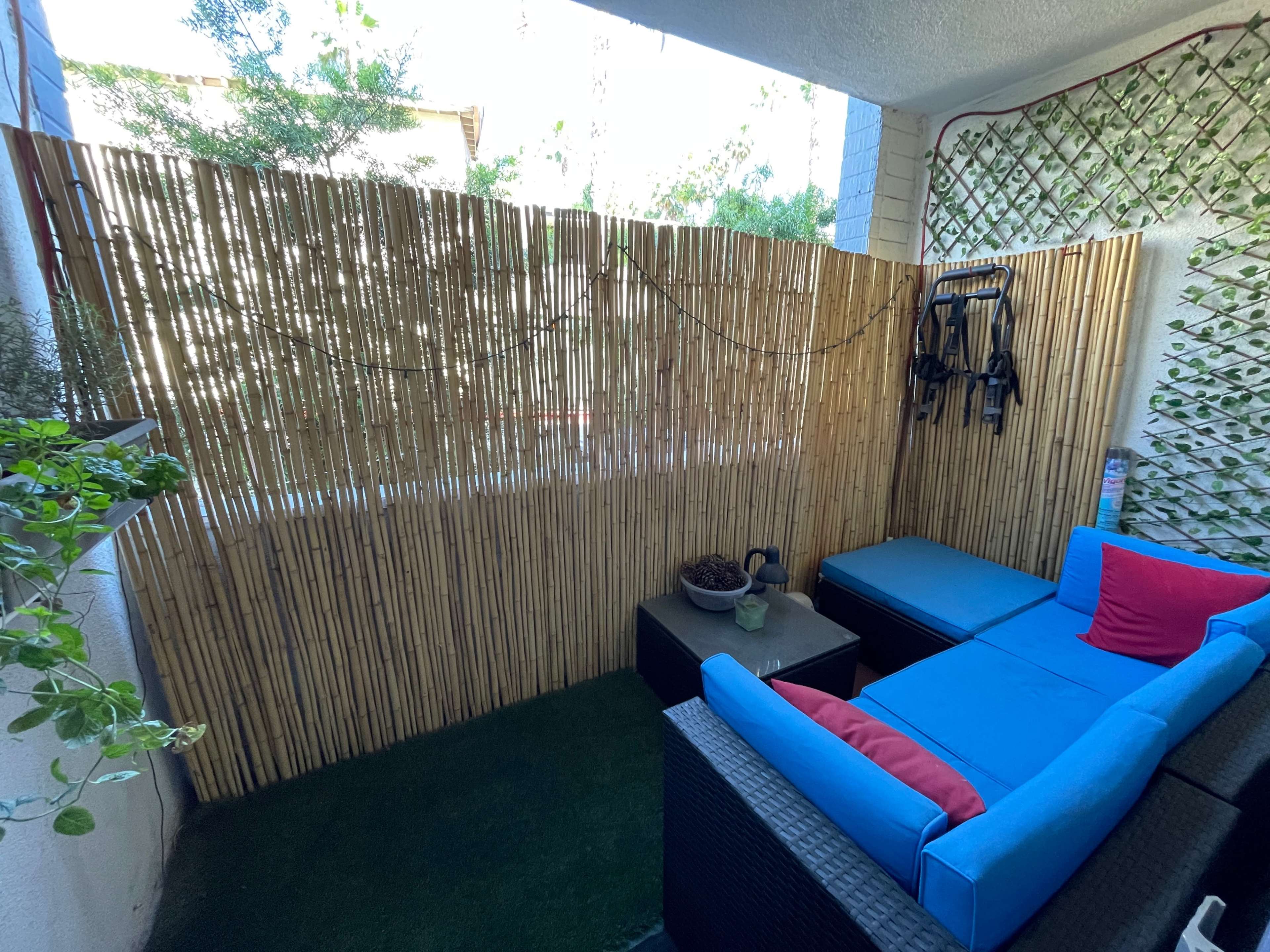 A small outdoor space enclosed by a bamboo fence, featuring a blue couch with red pillows, a coffee table, and potted plants.