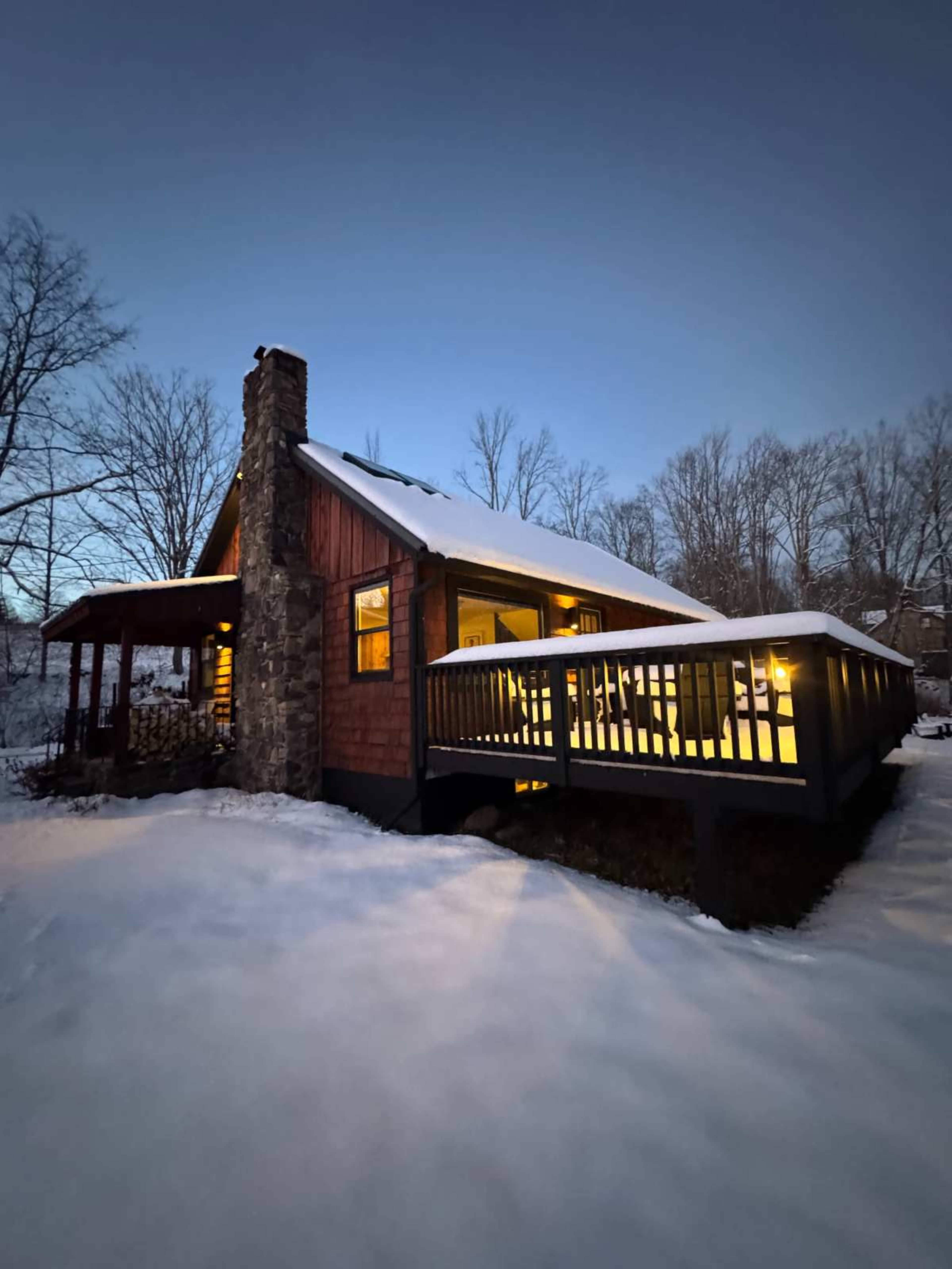 A cozy cabin with a snow-covered roof and a wooden deck is illuminated in the twilight.