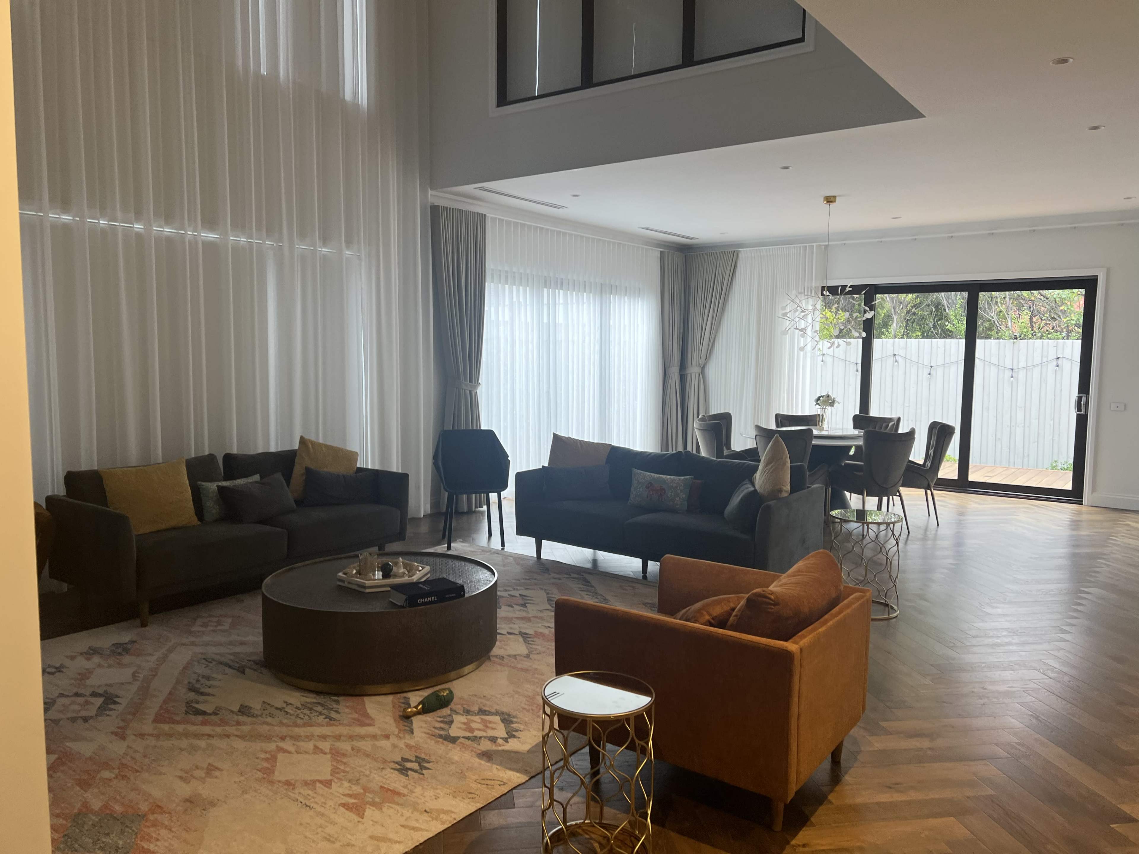 A spacious living room with large windows, featuring two dark sofas, a brown armchair, a round table with chairs, and a patterned rug on a wooden floor.