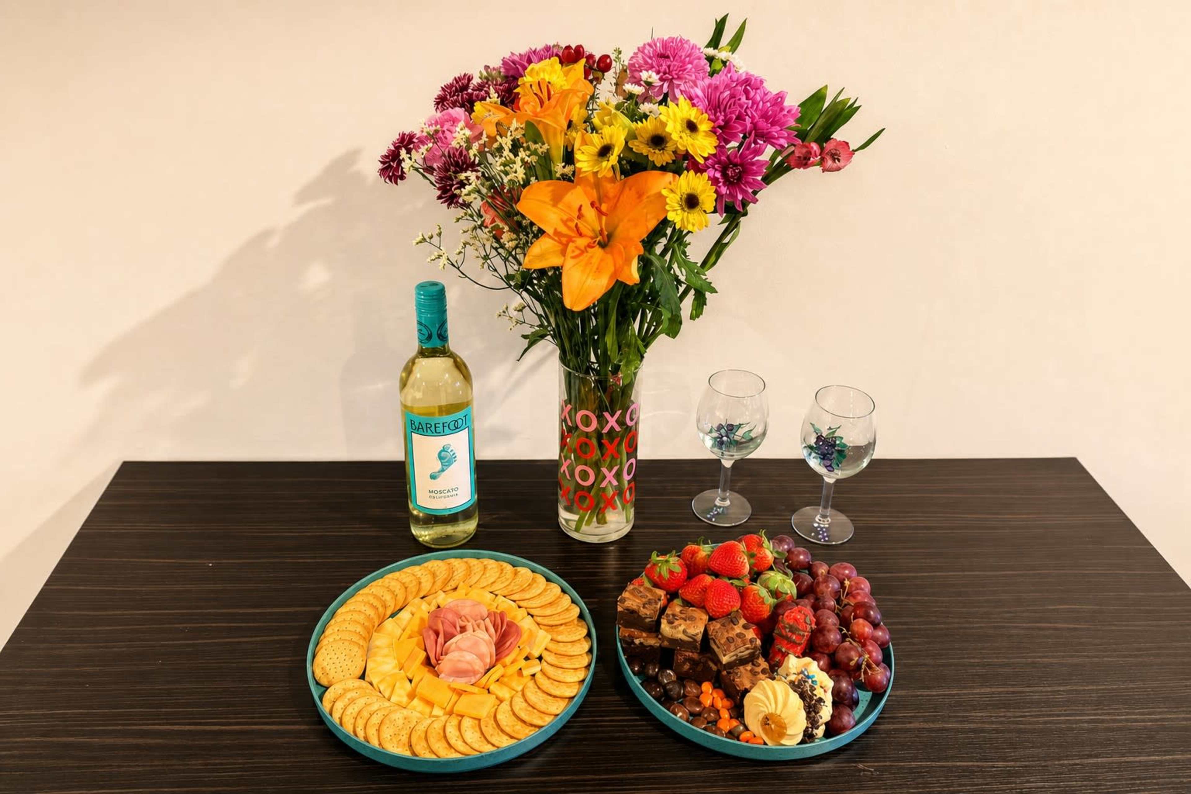 A table is set with a bottle of white wine, two decorative glasses, a vase of colorful flowers, and two plates filled with assorted fruits, snacks, and baked goods.