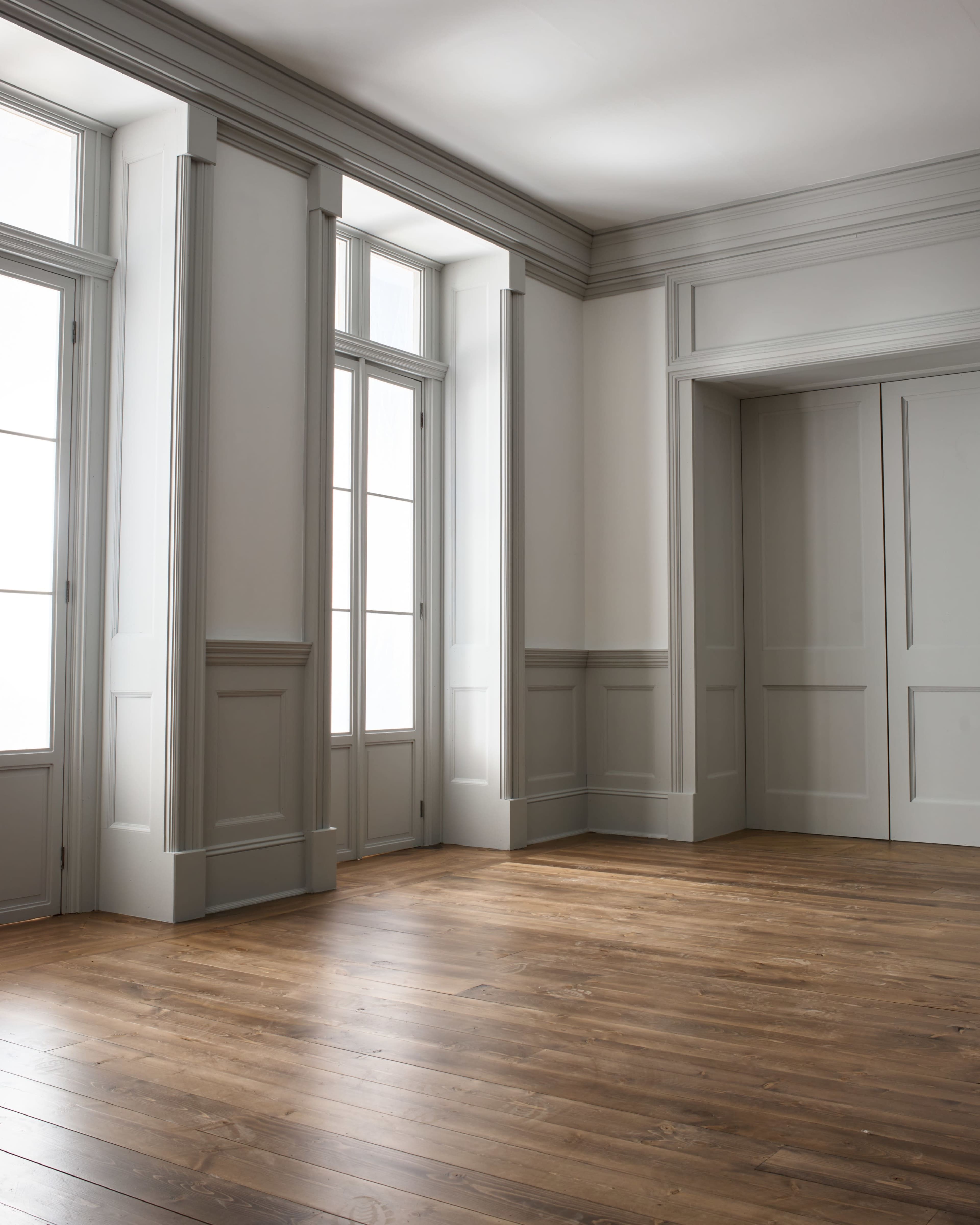 The image shows a partially empty room with wooden flooring, light gray walls, and large windows along two sides.