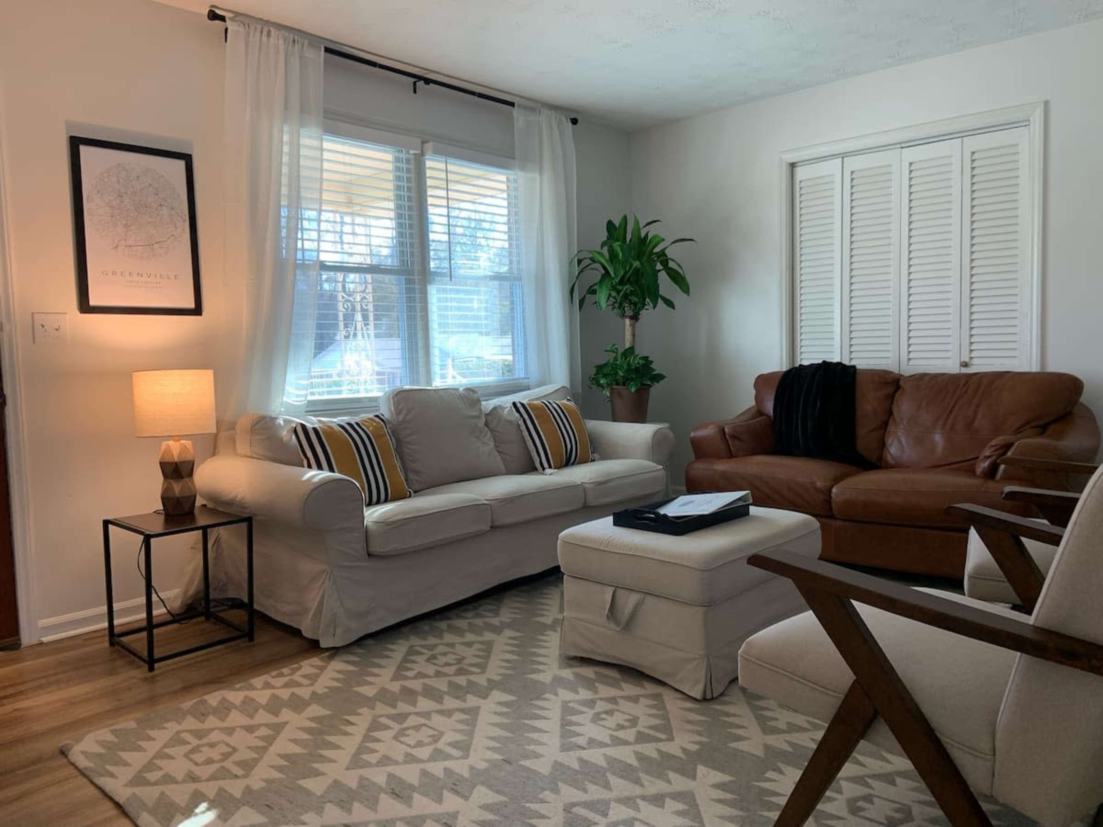 The living room features a light-colored couch and a brown leather sofa, with a small side table and a plant placed near the window.