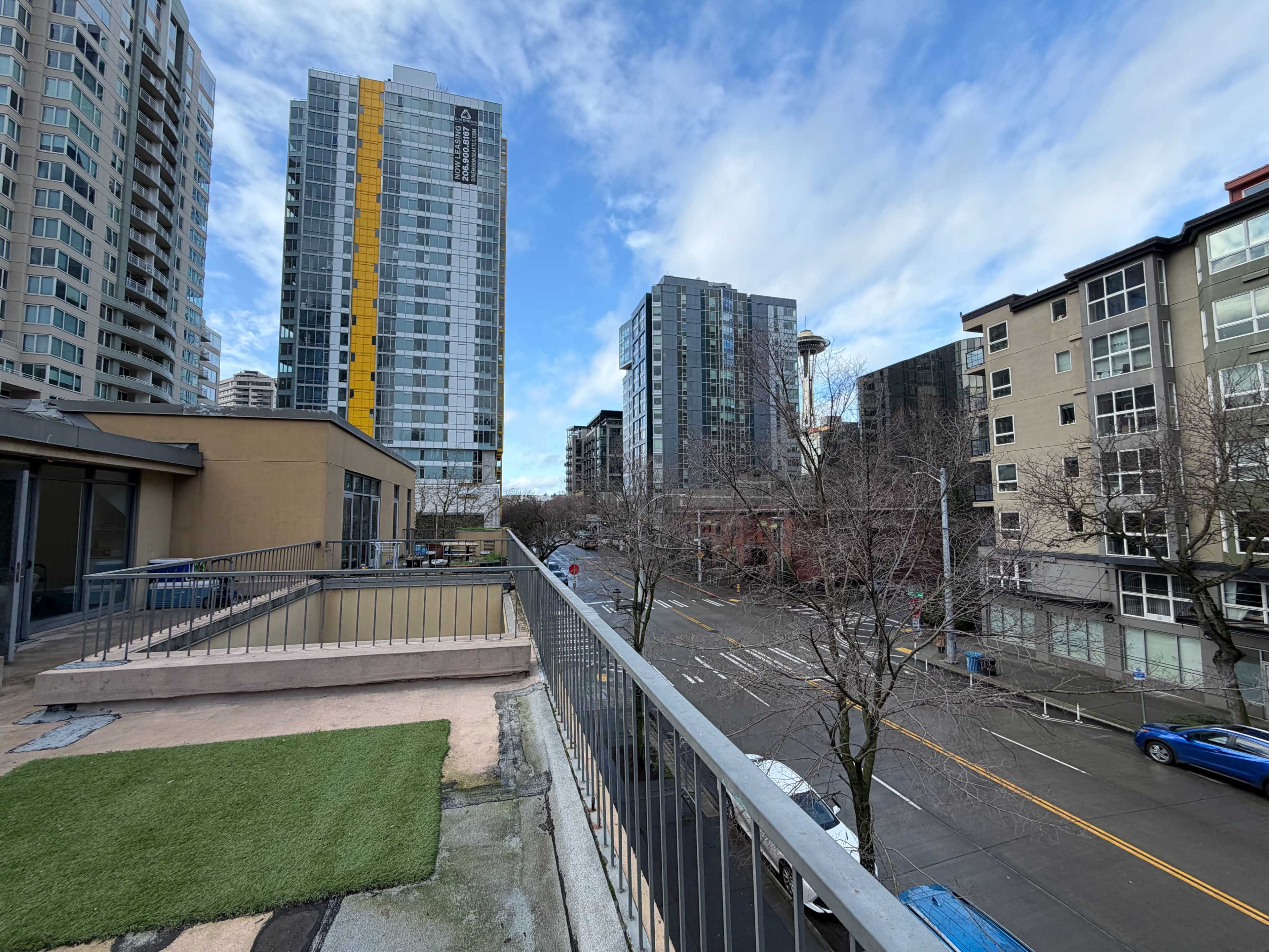 The image shows a view from a balcony overlooking a city street lined with modern buildings under a partly cloudy sky.