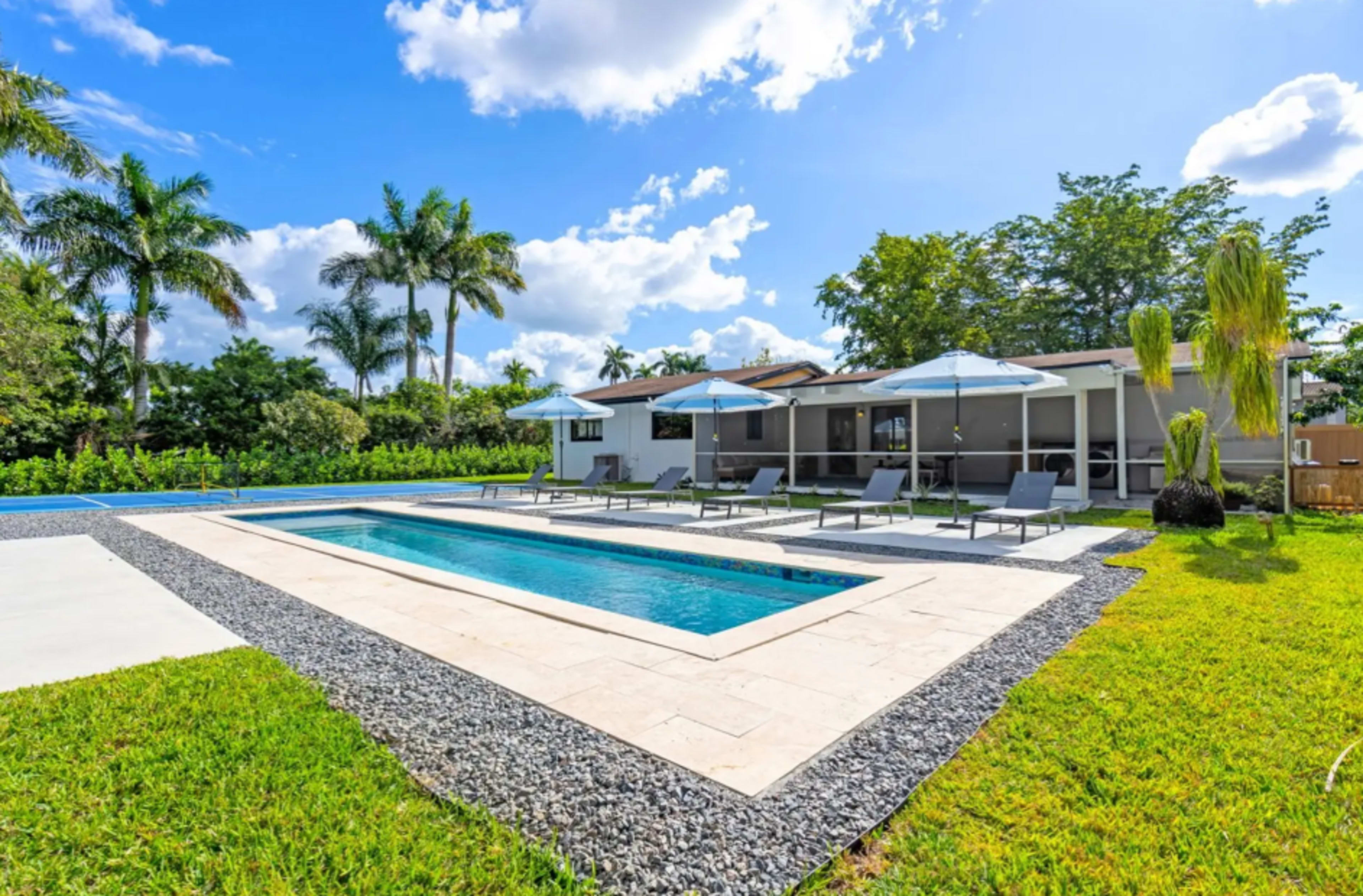 The image depicts a modern house with a swimming pool surrounded by lounge chairs and umbrellas, set amidst a lush green lawn and palm trees.