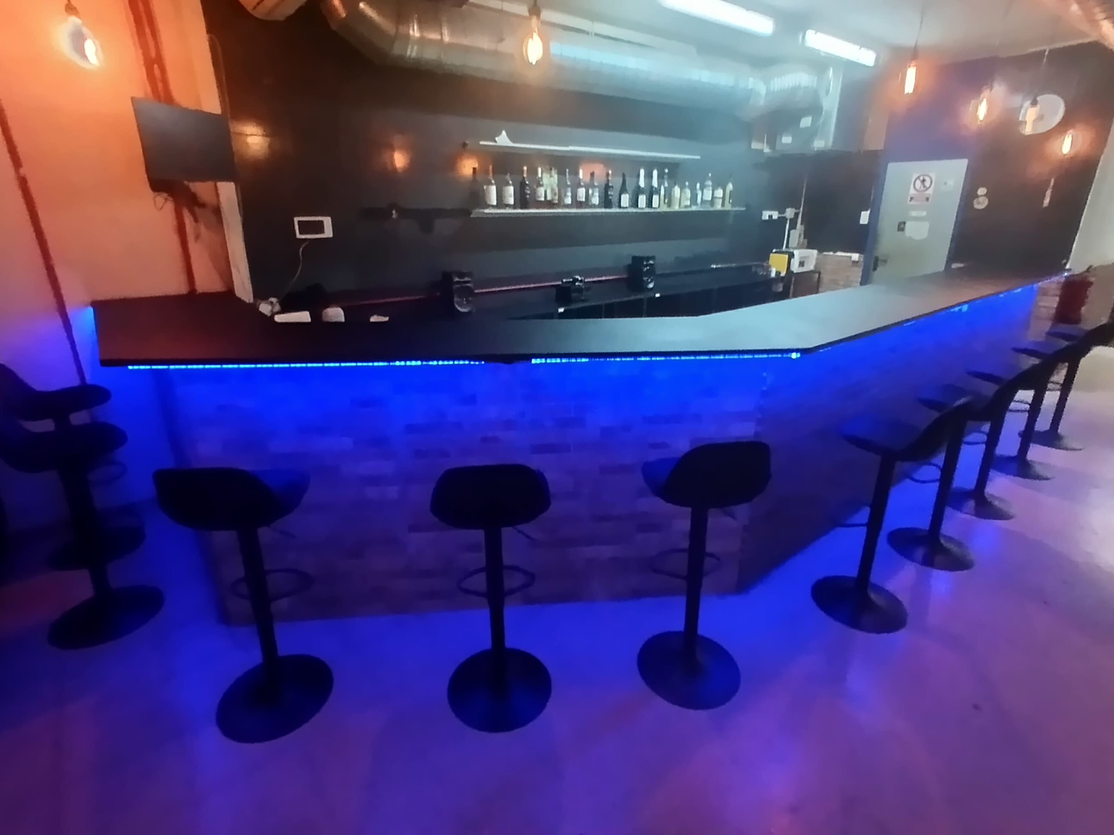 The image shows a modern bar area featuring a brick counter with blue LED lighting and several black stools arranged in front of it.