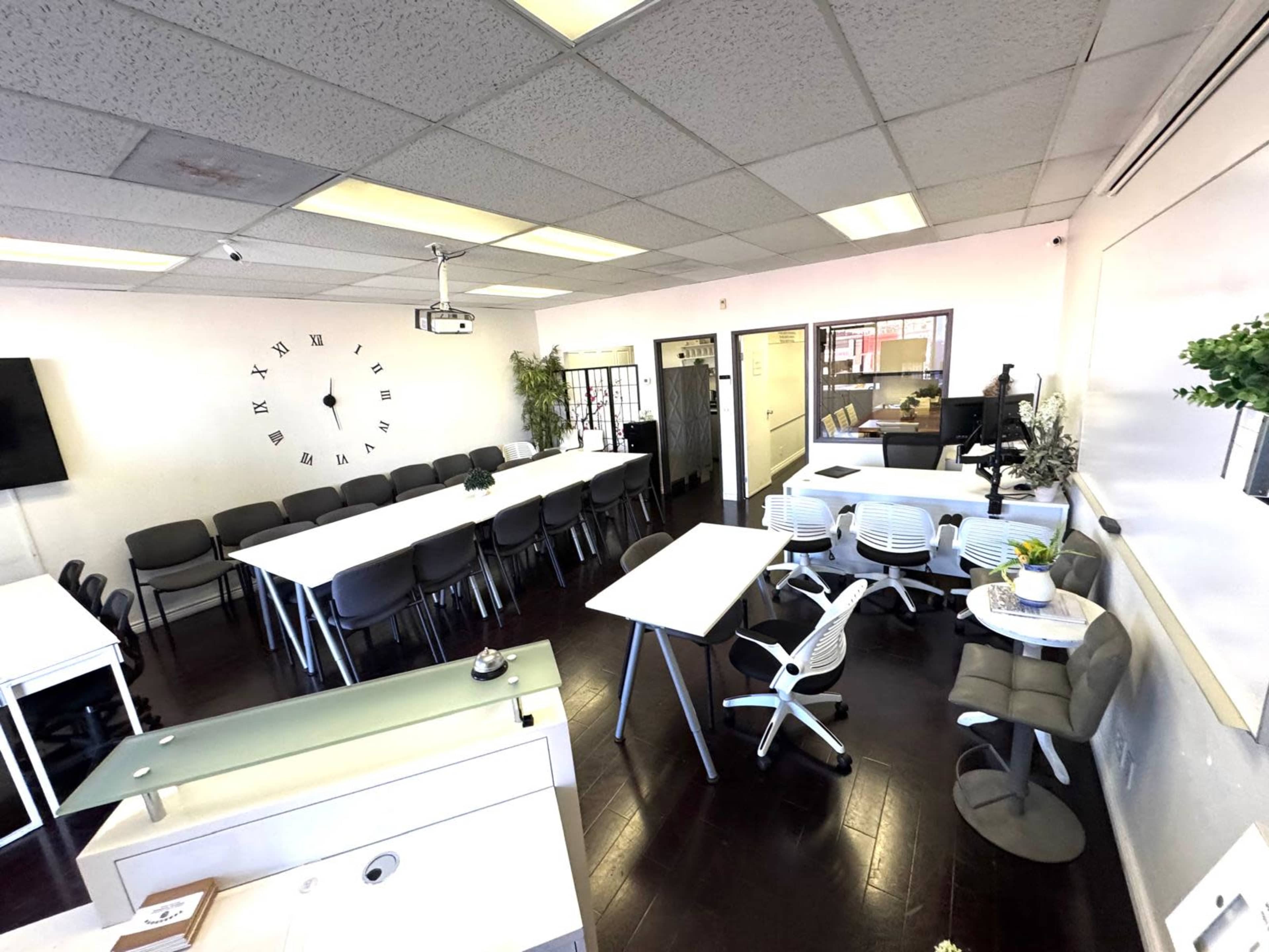 The image shows a bright office space featuring multiple tables and chairs arranged for meetings, with a clock on the wall and a desk in the corner.