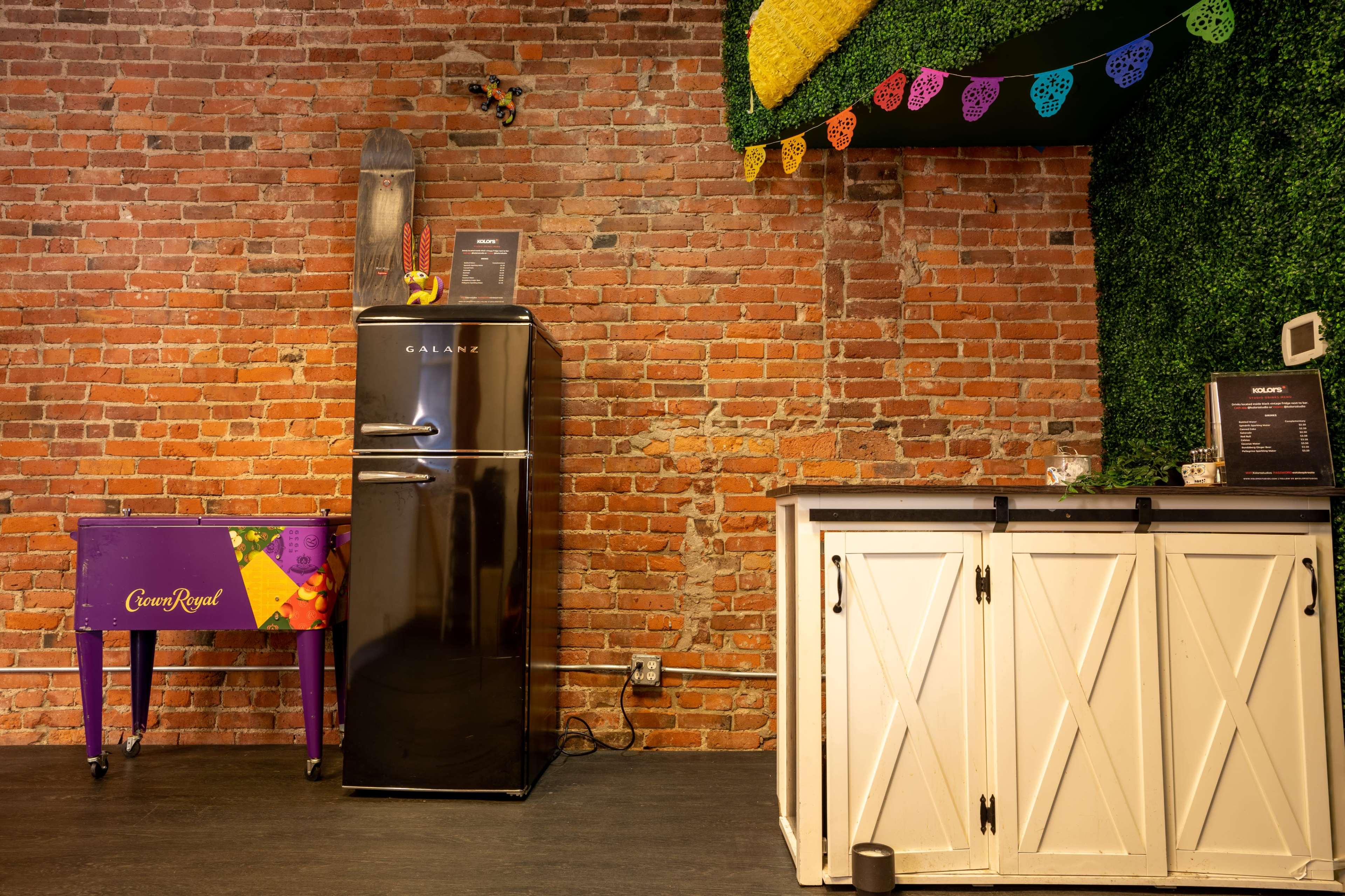 The scene features a black fridge next to a purple table with a Crown Royal logo, set against a backdrop of exposed brick walls and decorative foliage.