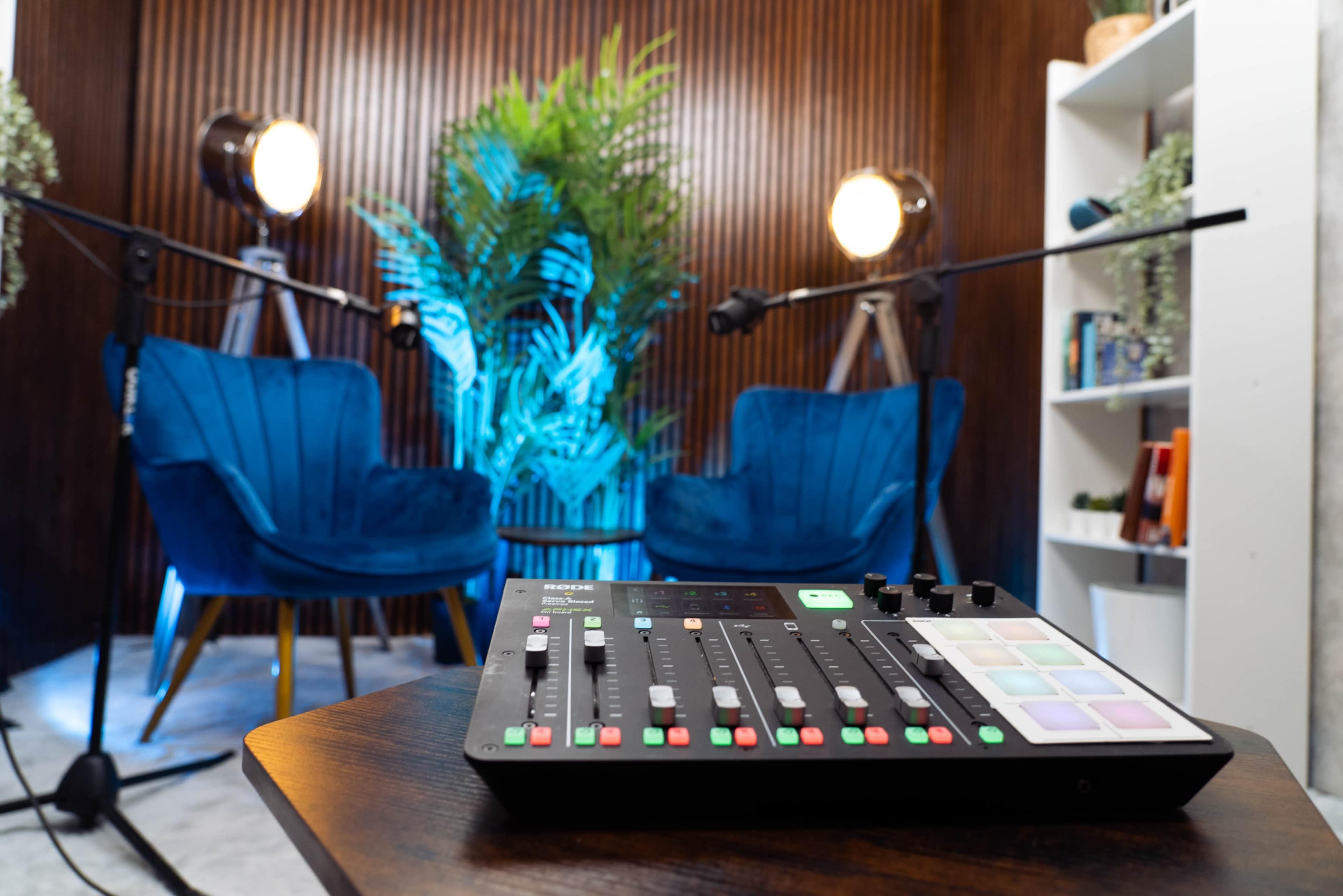 A sound mixing console in the foreground, with two blue chairs and studio lights in a modern recording studio setup behind it.