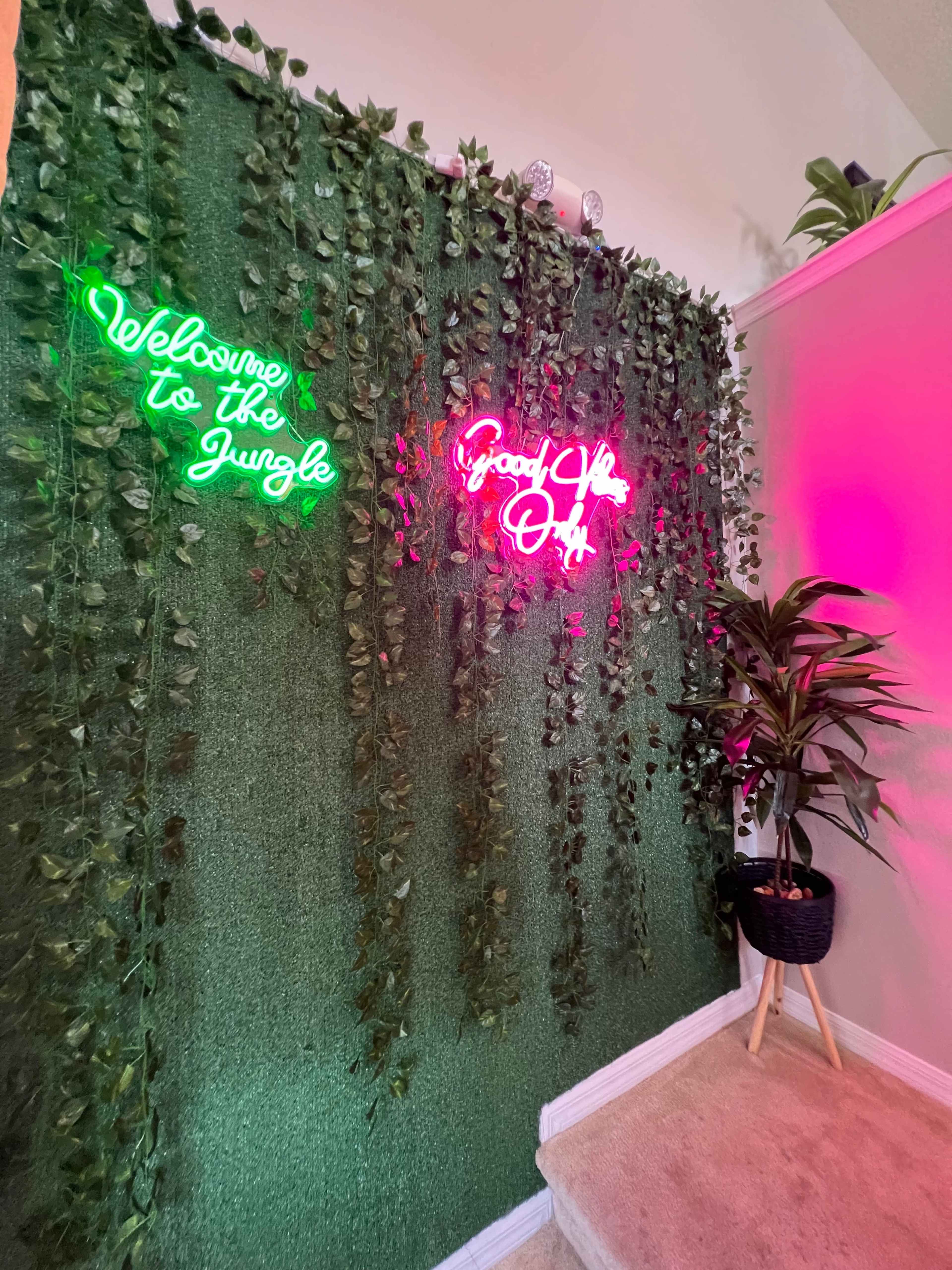 The image shows a wall covered in greenery with neon signs that read "Welcome to the Jungle" and "Good Vibes Only," alongside a plant in a basket on a stand.