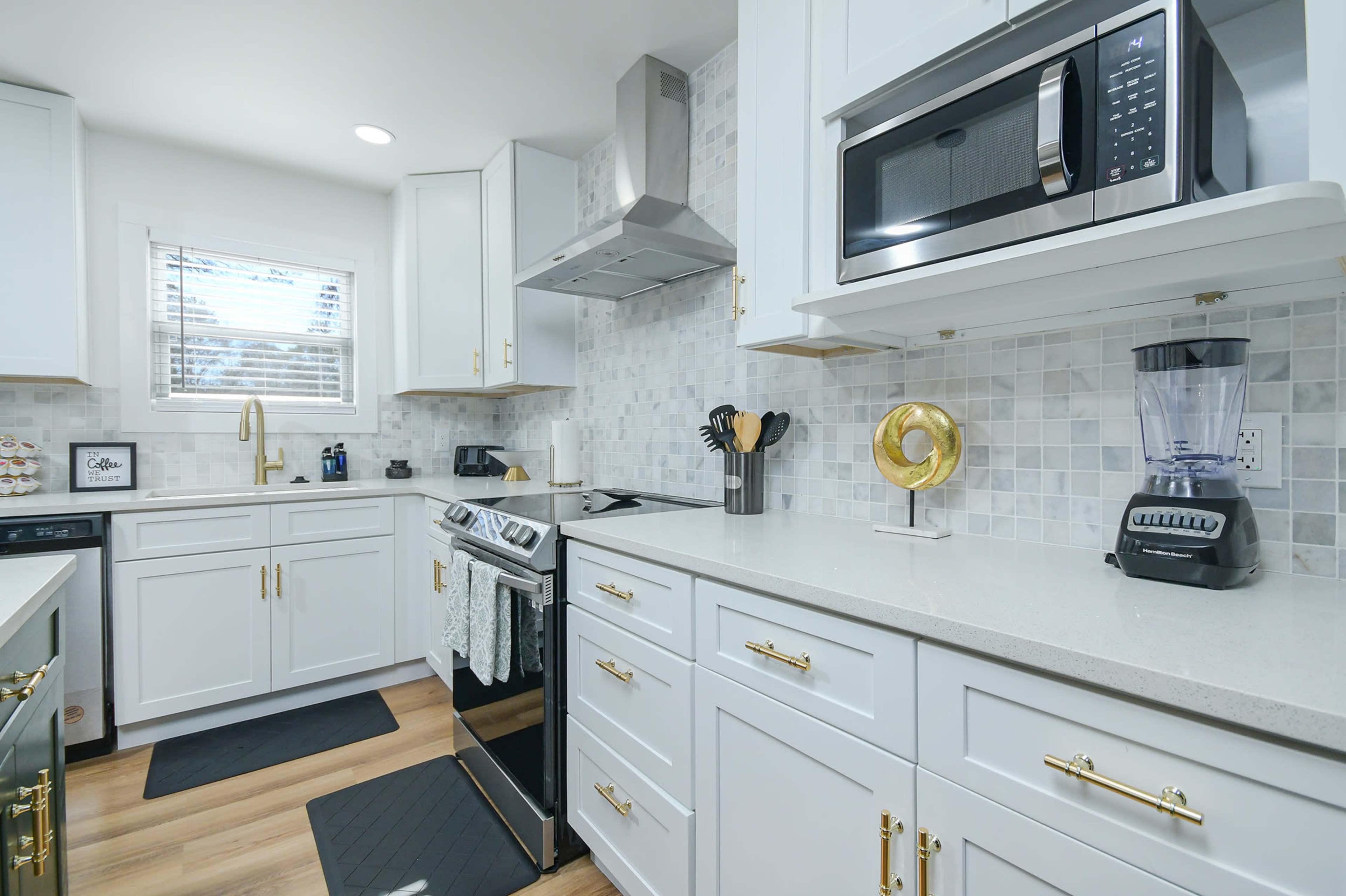 The kitchen features white cabinetry, a stainless steel microwave, and a blender next to a stove on a light countertop with a mosaic tile backsplash.