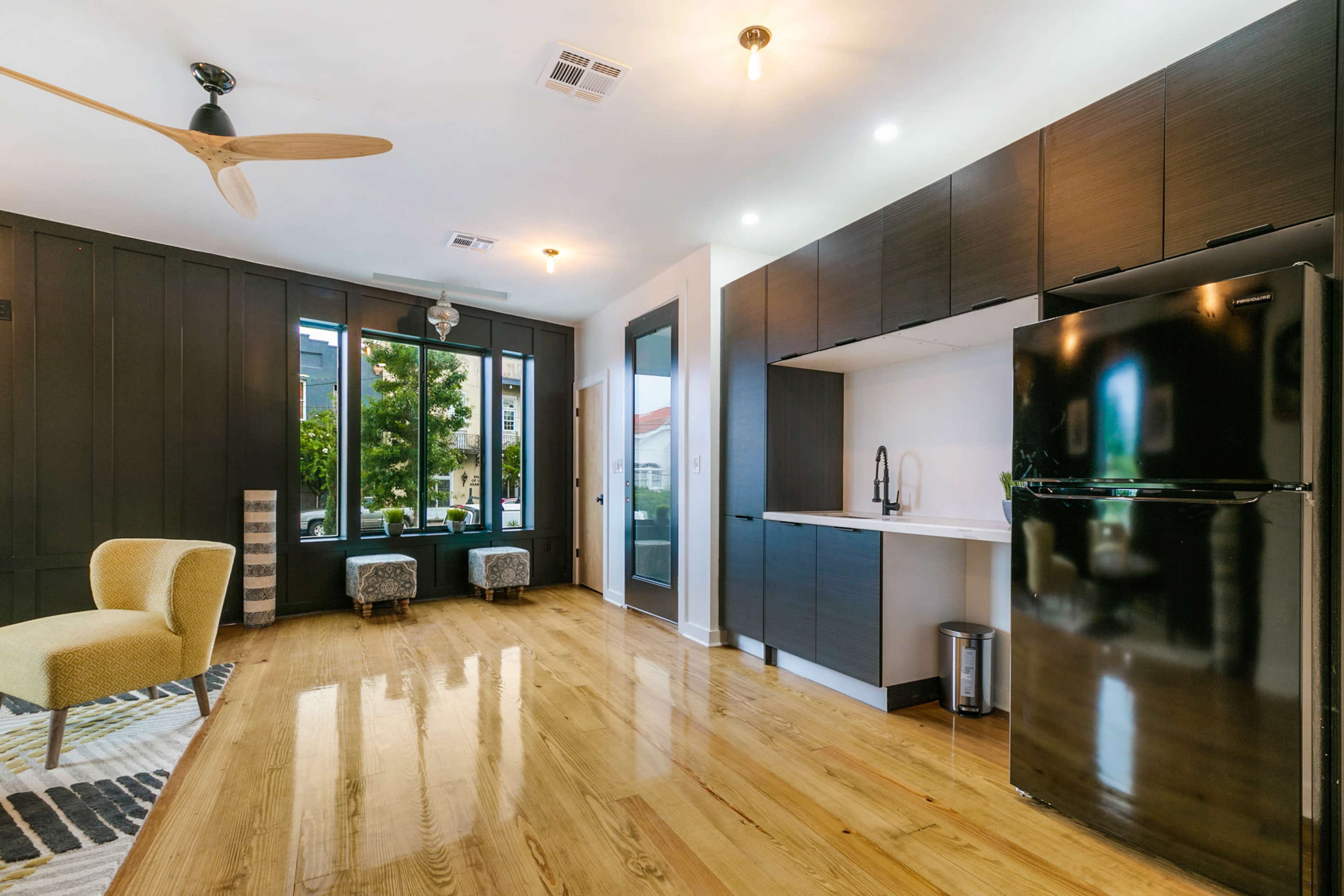 A modern kitchen and living area featuring dark cabinetry, a black refrigerator, a wooden floor, and large windows with greenery outside.