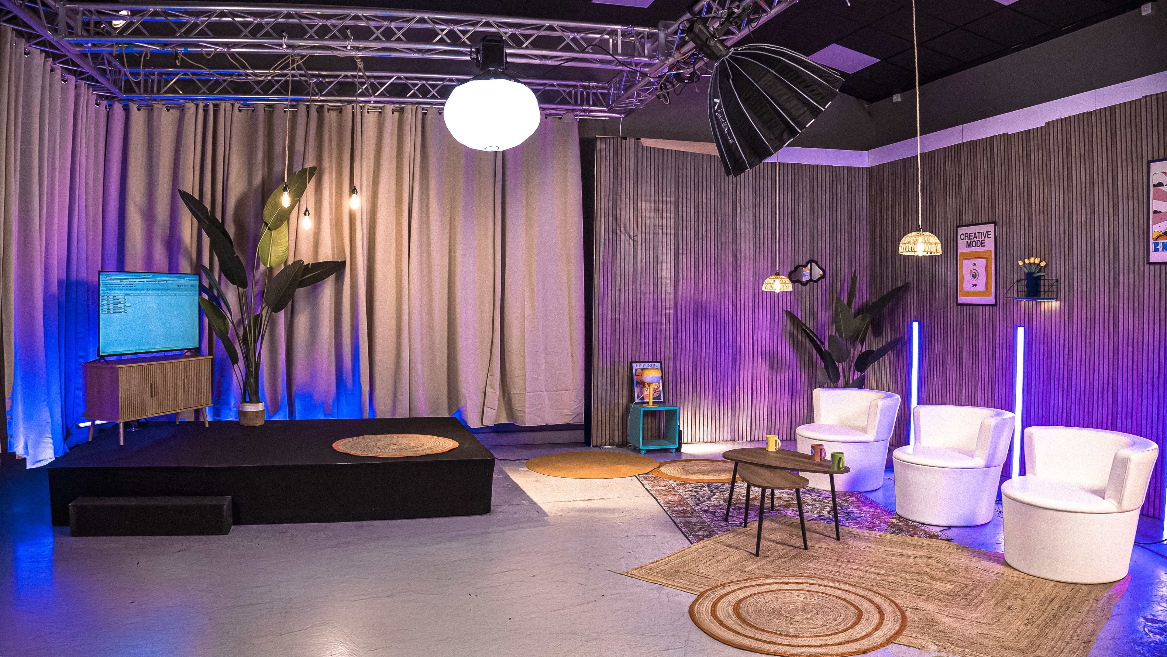 The image shows a modern presentation studio with a stage, white chairs, a television, and decorative plants, illuminated by soft lighting.