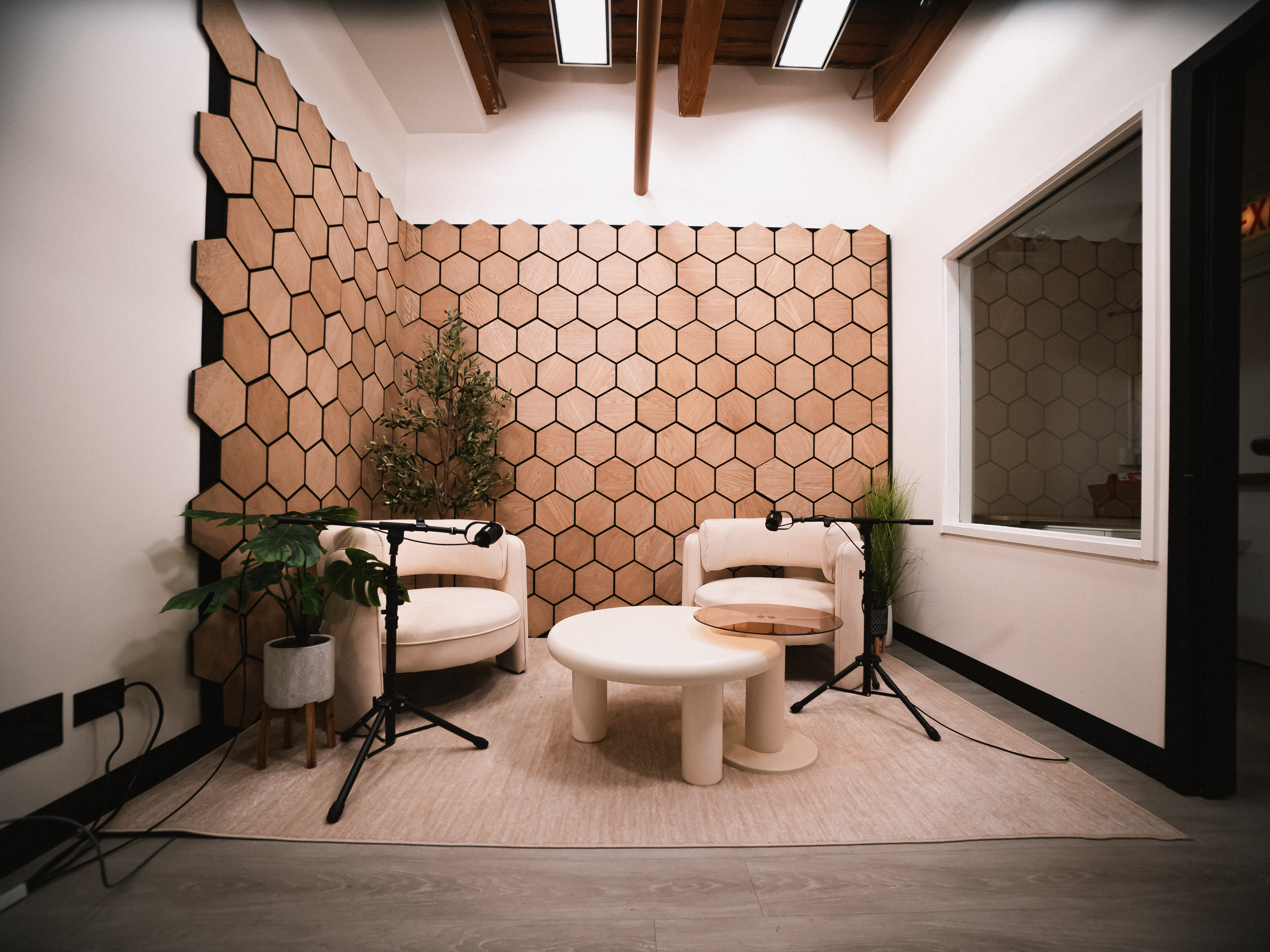 The image shows a cozy podcasting studio featuring two white armchairs, a circular coffee table, and a hexagonal wooden accent wall, with plants and microphones present.