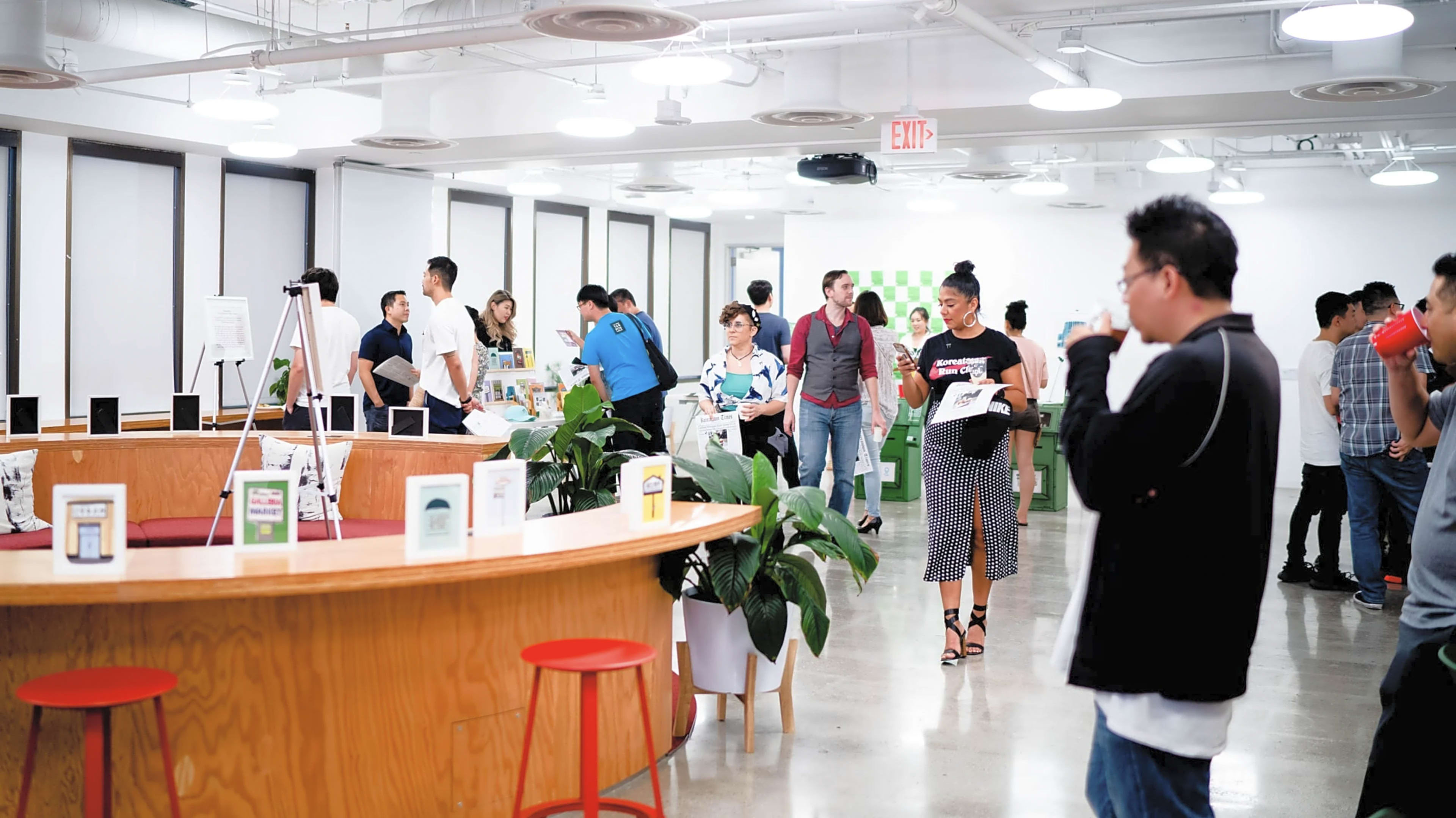 A group of people engages in conversation and activities in a modern workspace featuring a central wooden counter and greenery.