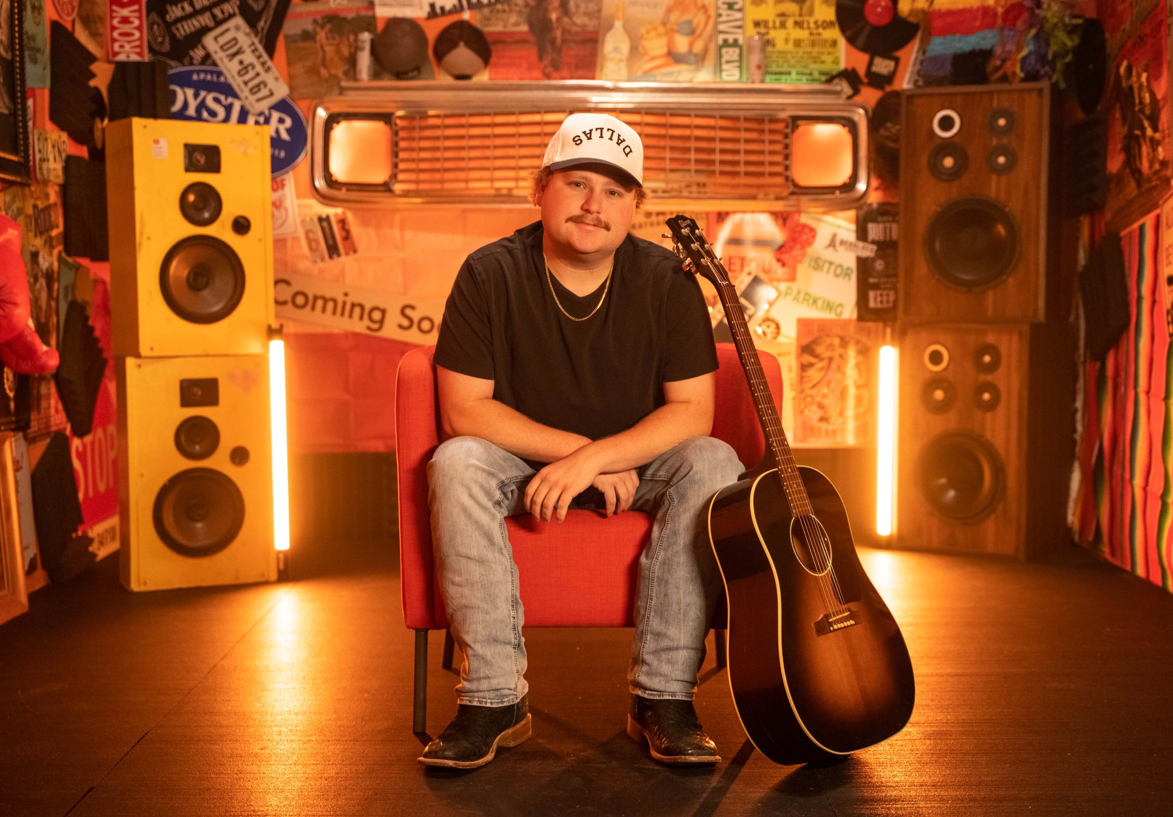 A musician sits in a red chair with a guitar beside him, surrounded by colorful posters and speakers in a vibrant studio setting.