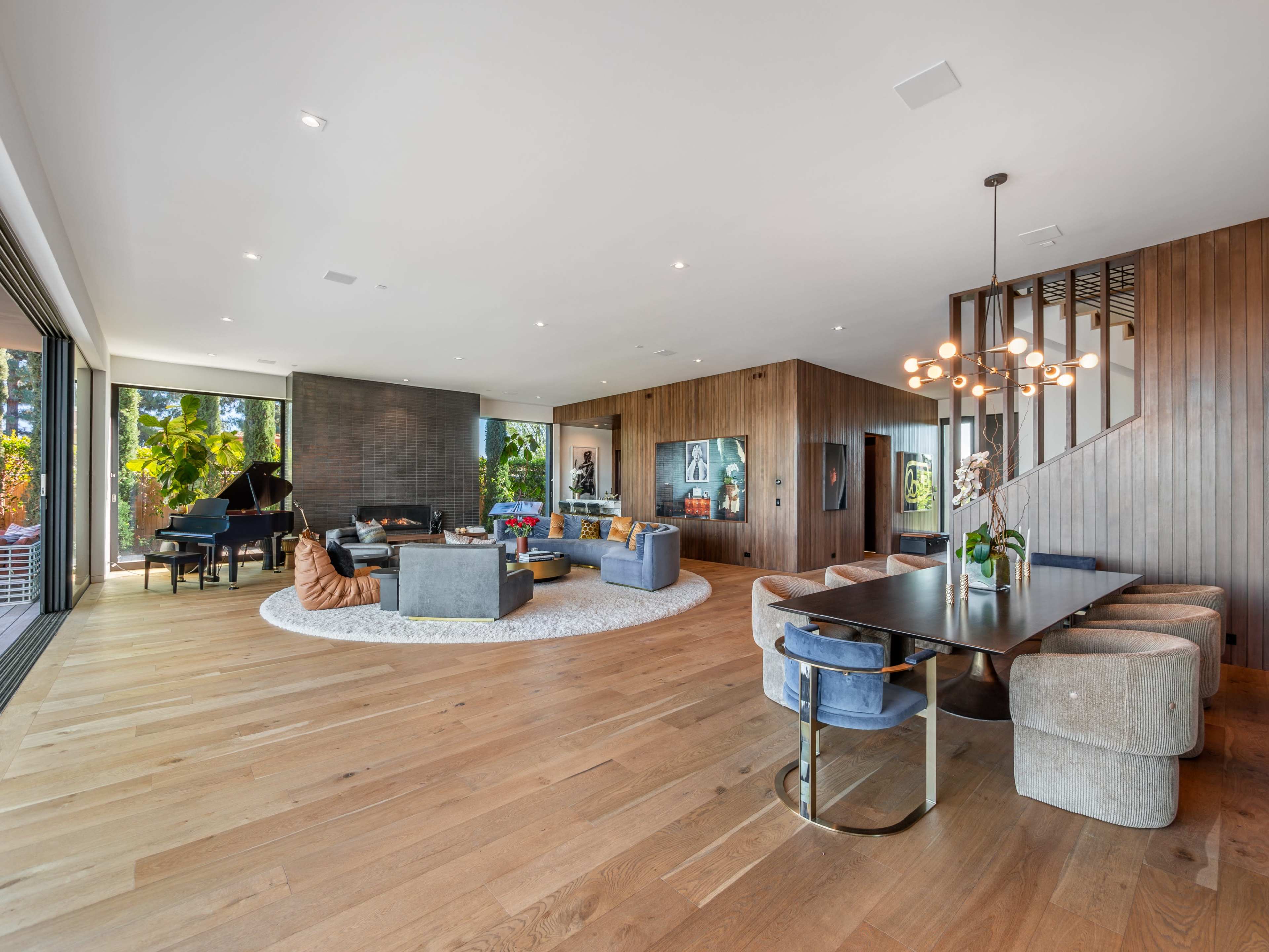 A modern open-plan living and dining area with large windows, wood accents, and a piano in one corner.