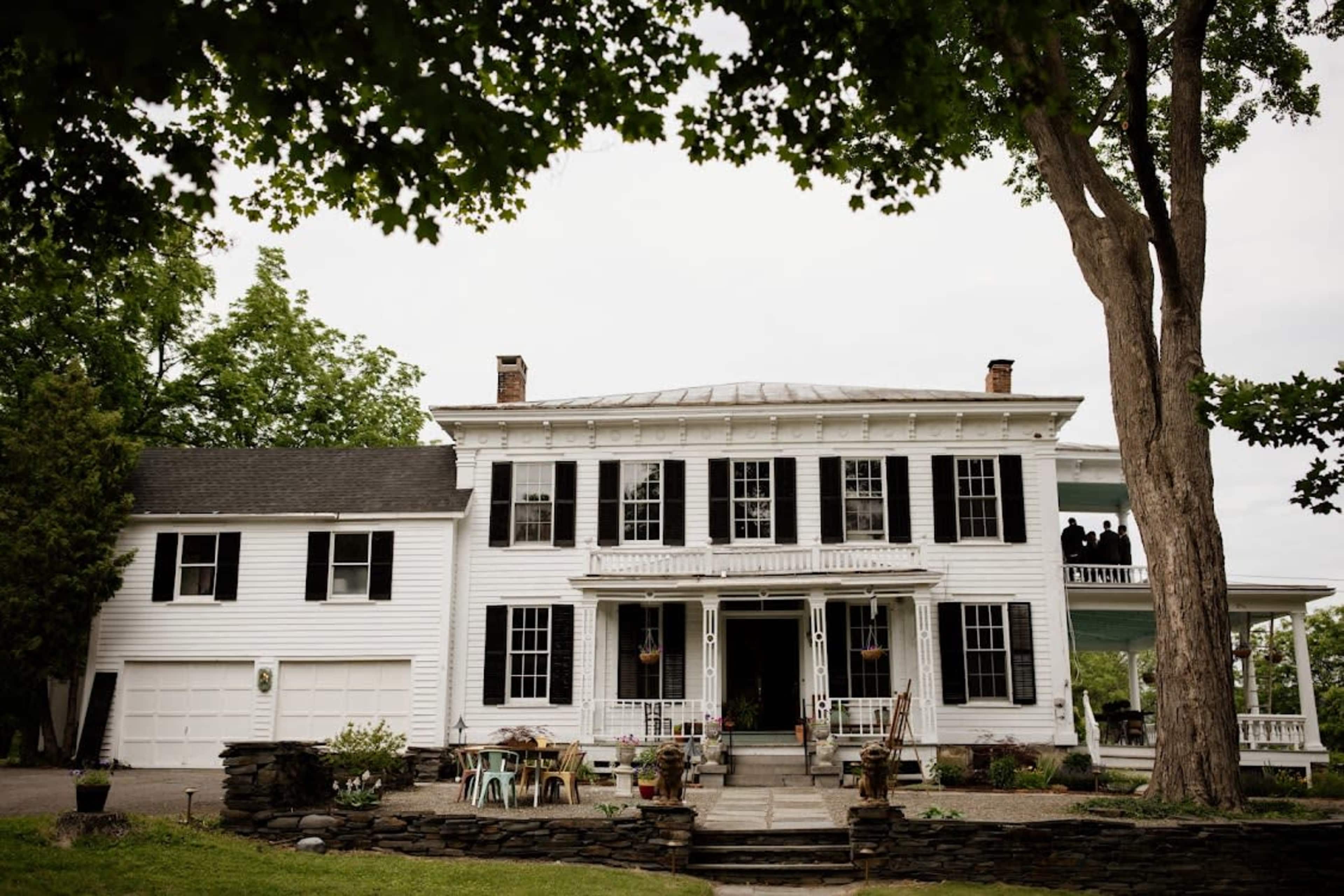 A large white house with black shutters, a central entrance, and a porch, surrounded by trees and patio furniture.