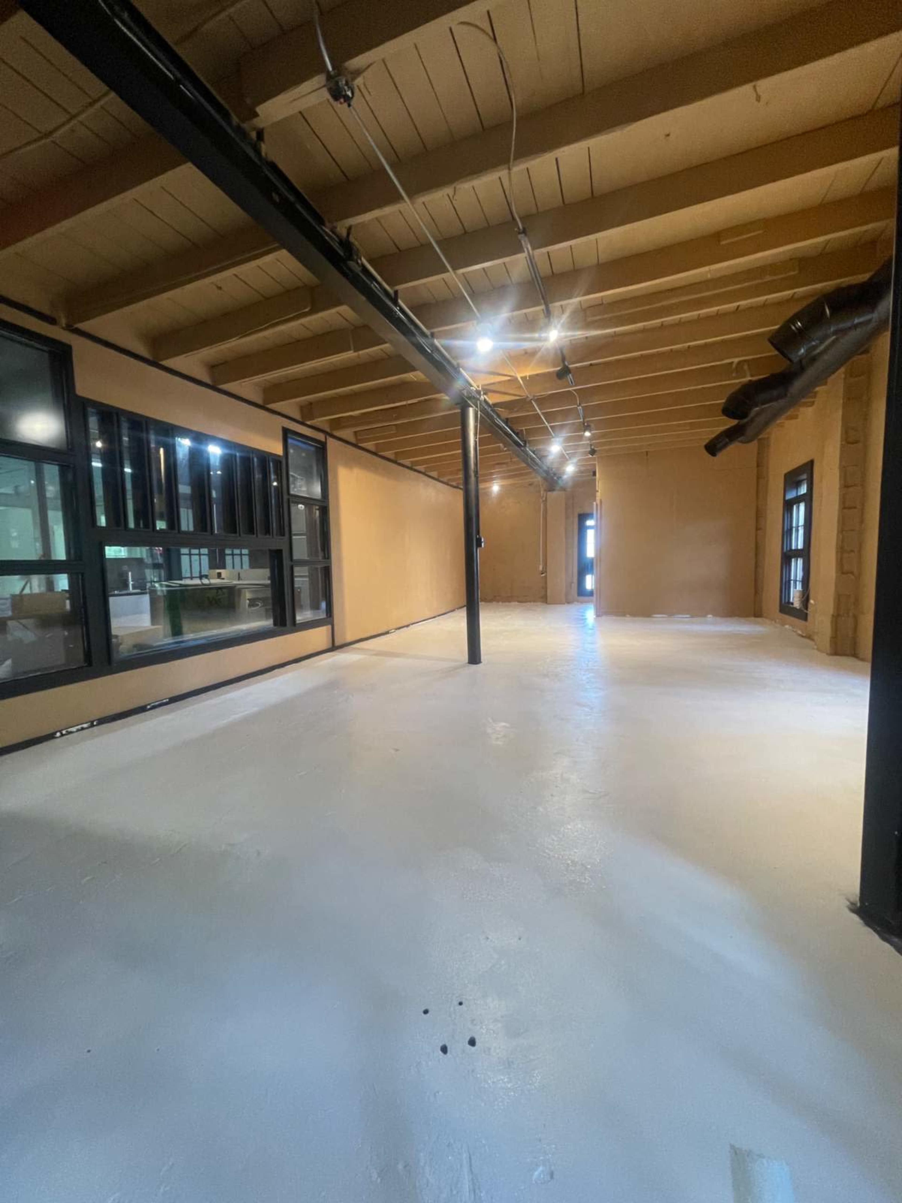 The image shows an unfinished interior space with a white floor, exposed beams on the ceiling, and large windows along one side.