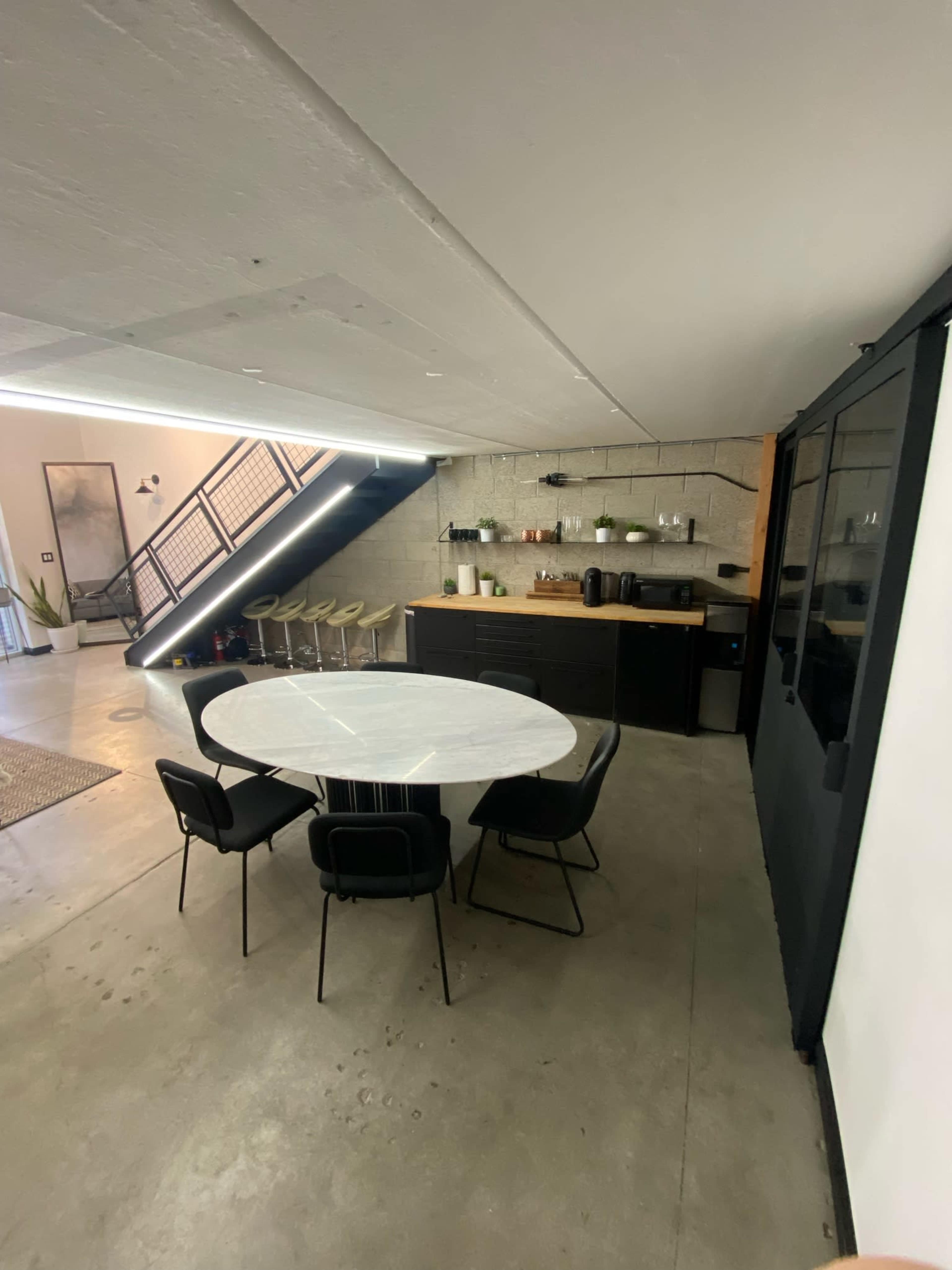 The image shows a modern dining area with a round white table surrounded by black chairs, adjacent to a kitchen area and a staircase.