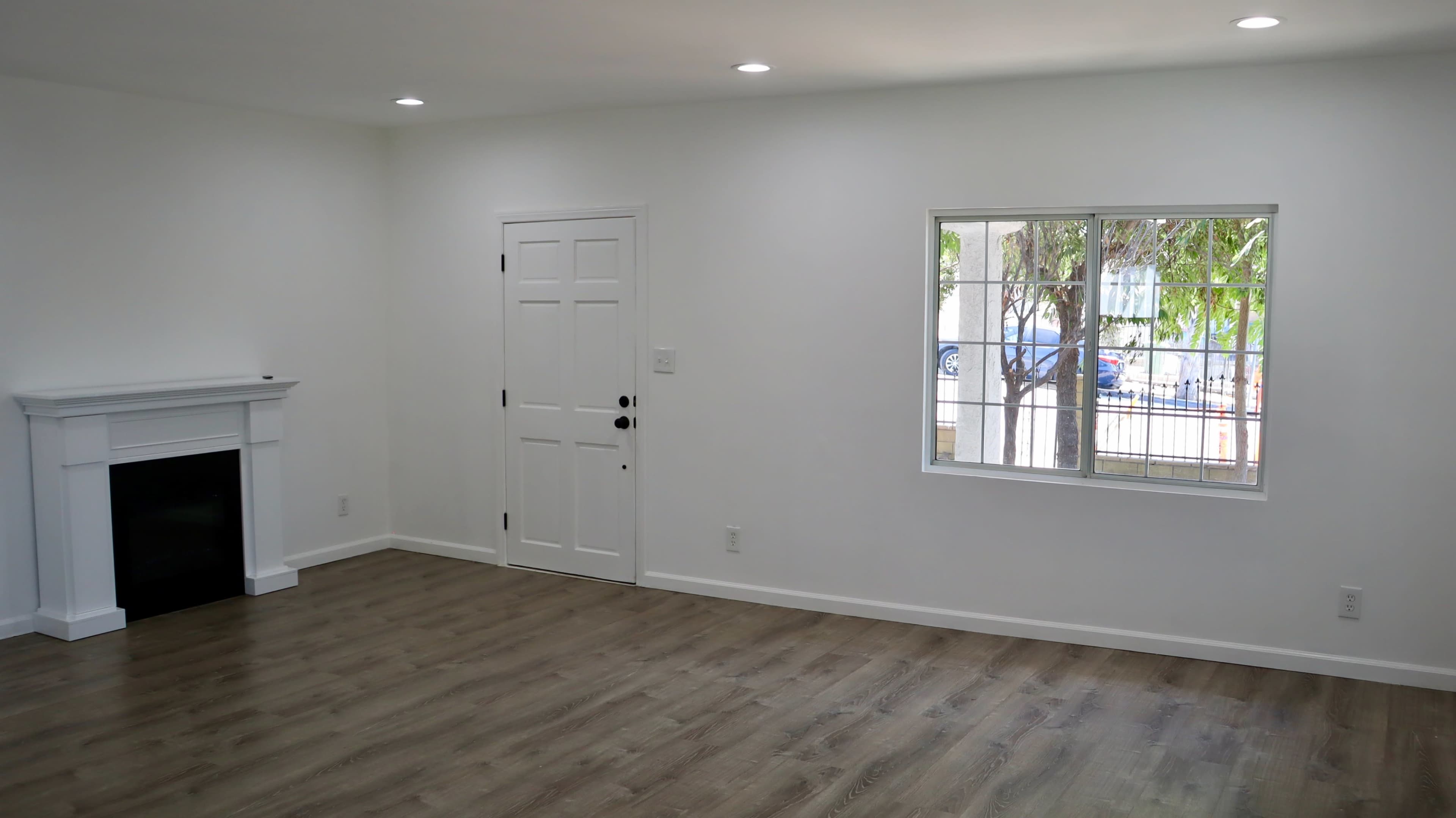 A bright, empty living room with a white wall, a door, a fireplace, and a window with trees visible outside.