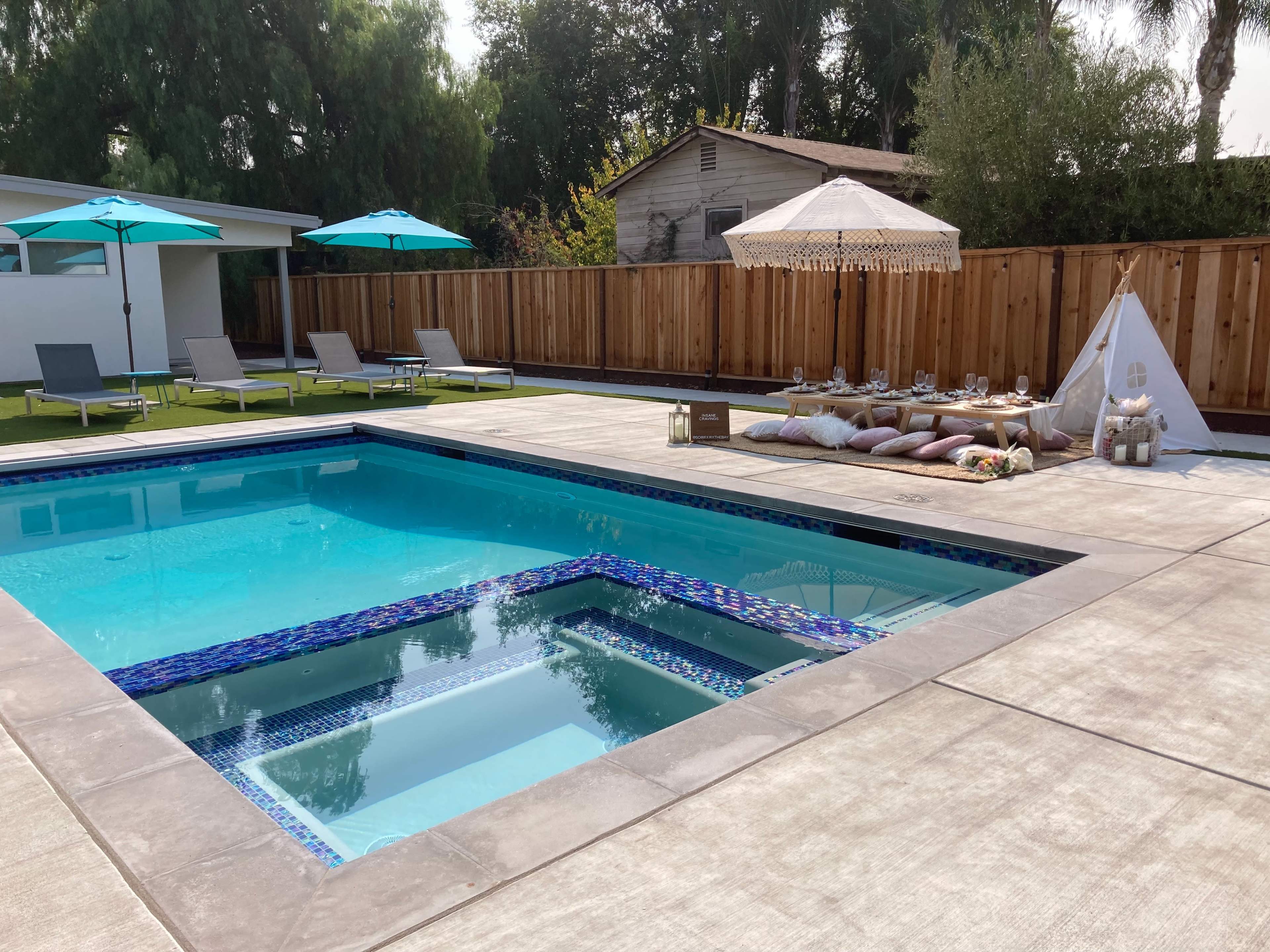 A backyard pool area with lounge chairs, blue umbrellas, and a covered seating arrangement beside the pool.
